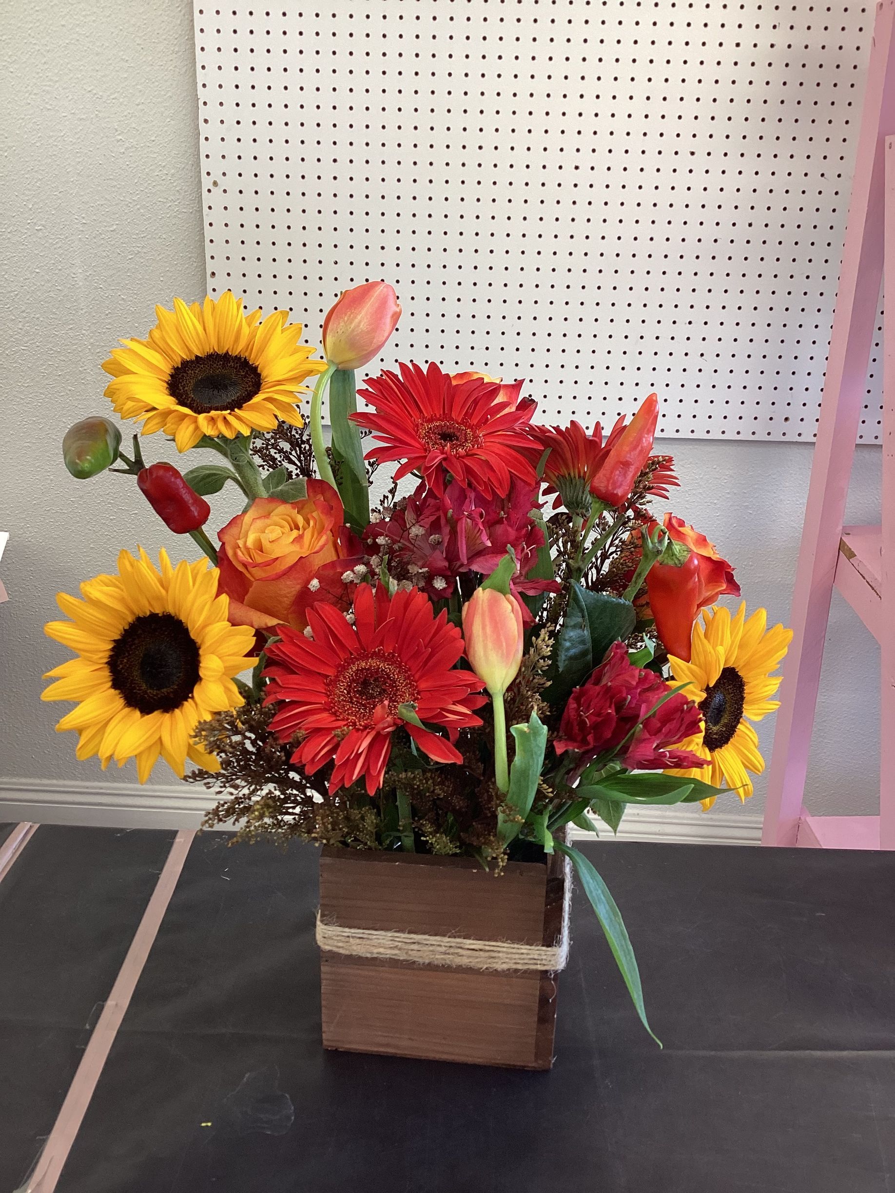 A wooden cube vase holds a vibrant floral arrangement of sunflowers, red gerbera daisies, orange roses, and peach tulips.