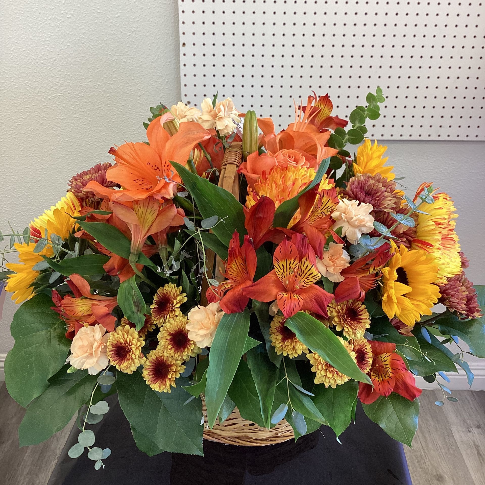 A lush autumn-themed flower arrangement with orange lilies, yellow sunflowers, and chrysanthemums in a wicker basket.