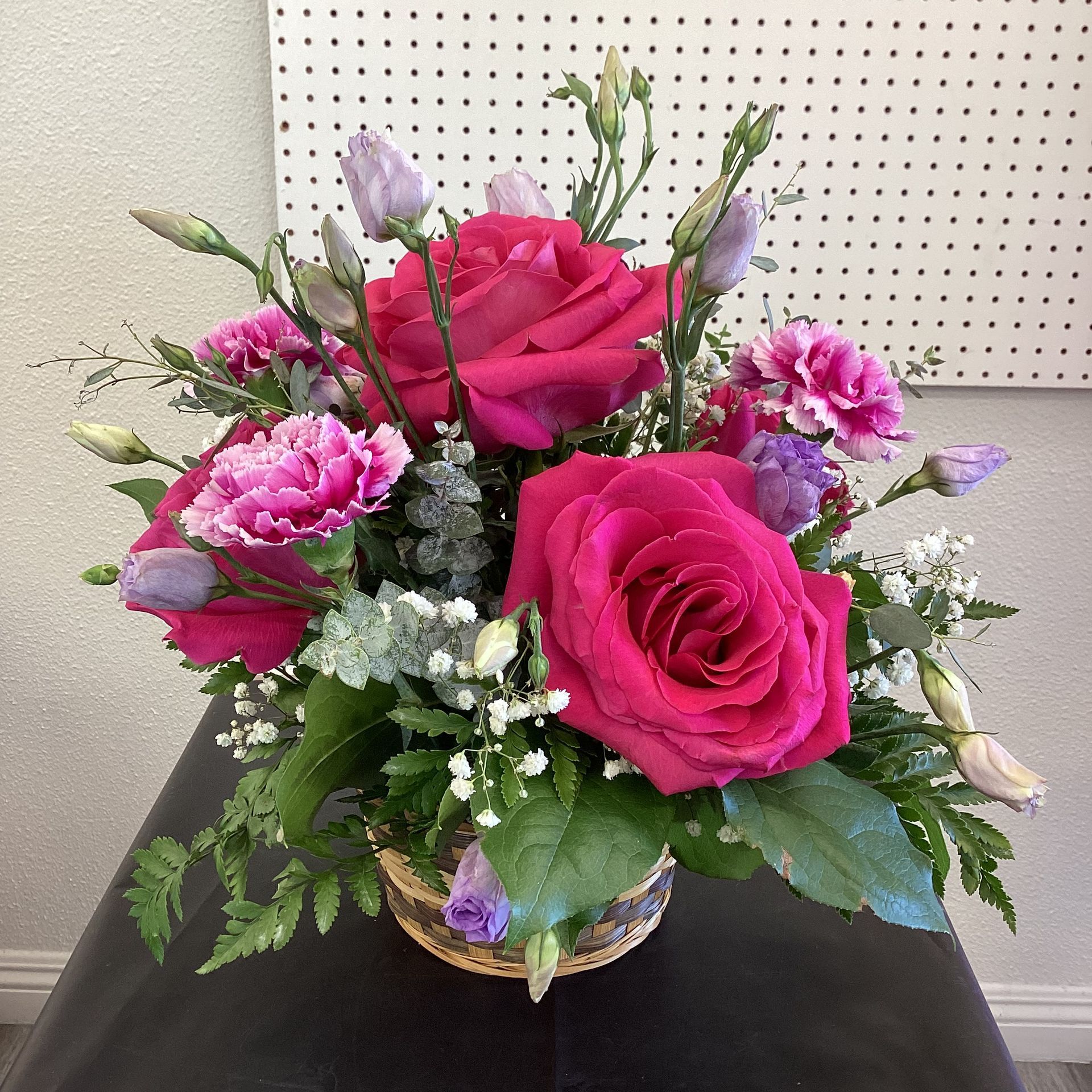 A vibrant bouquet featuring large dark pink roses, carnations, and light purple lisianthus buds in a woven basket.