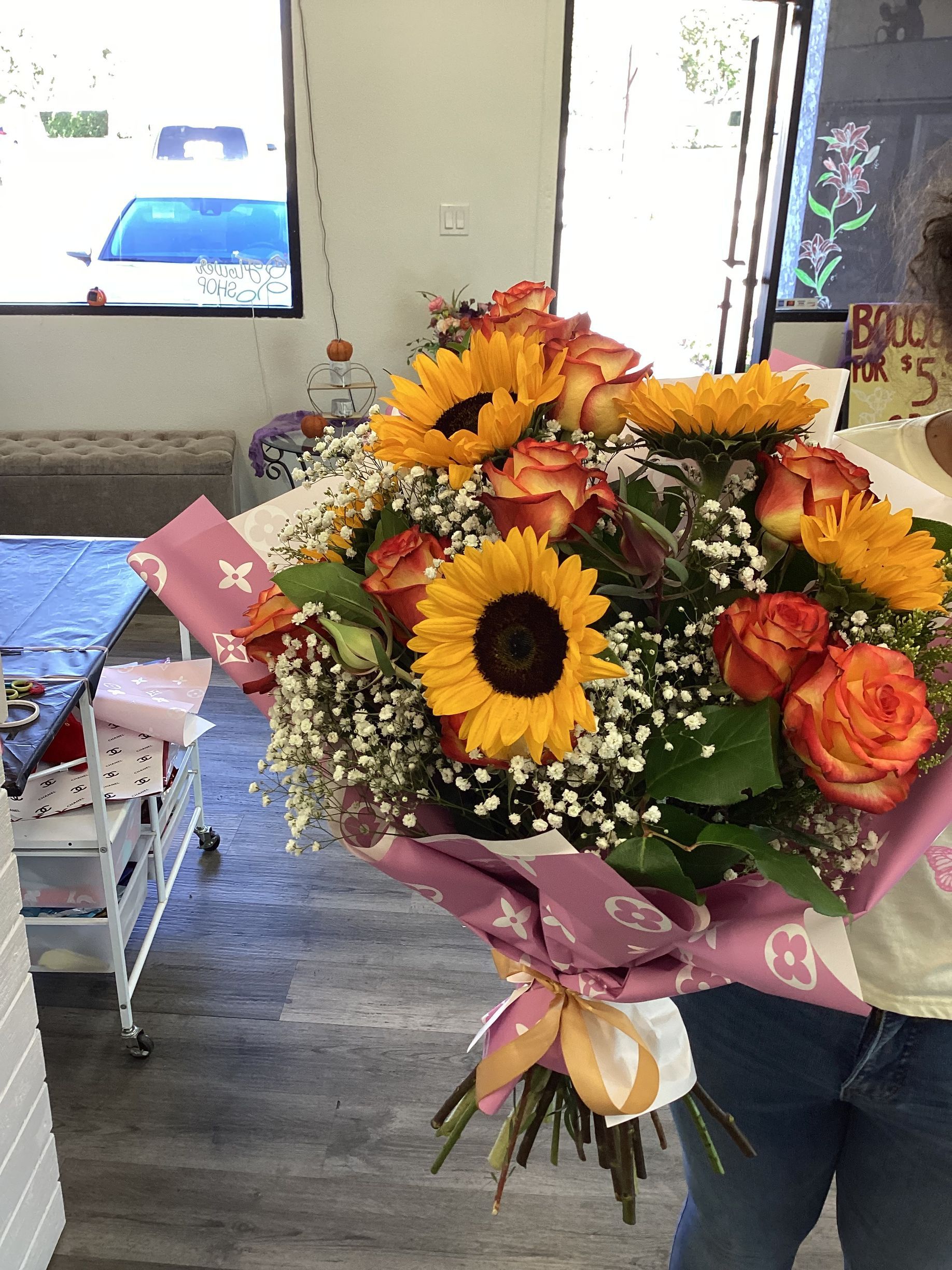 A person holds a bouquet of sunflowers and orange roses wrapped in pink floral paper against a bright shop background.
