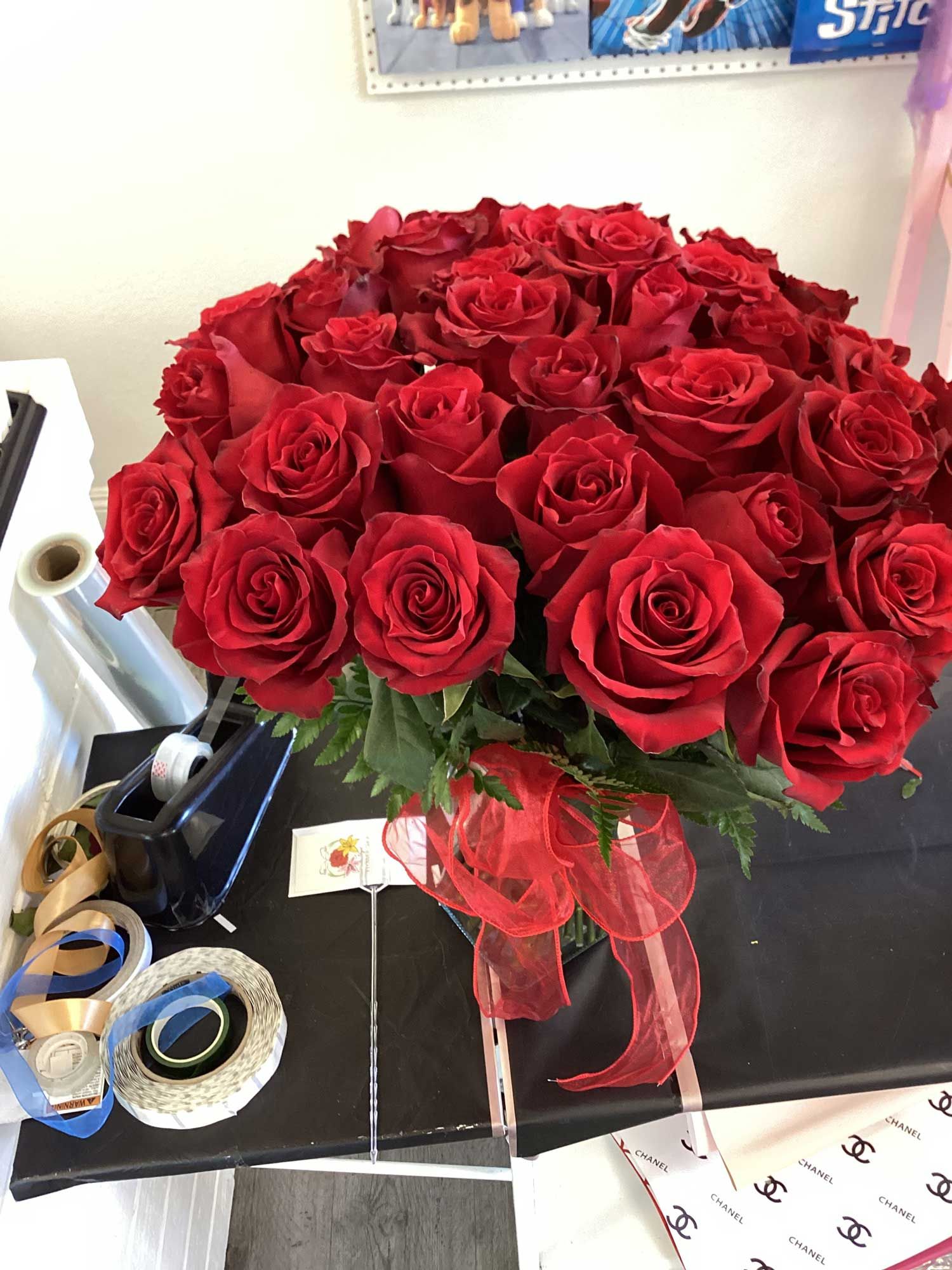 A large bouquet of vibrant red roses tied with a matching red ribbon sits on a desk next to tape and ribbon rolls.