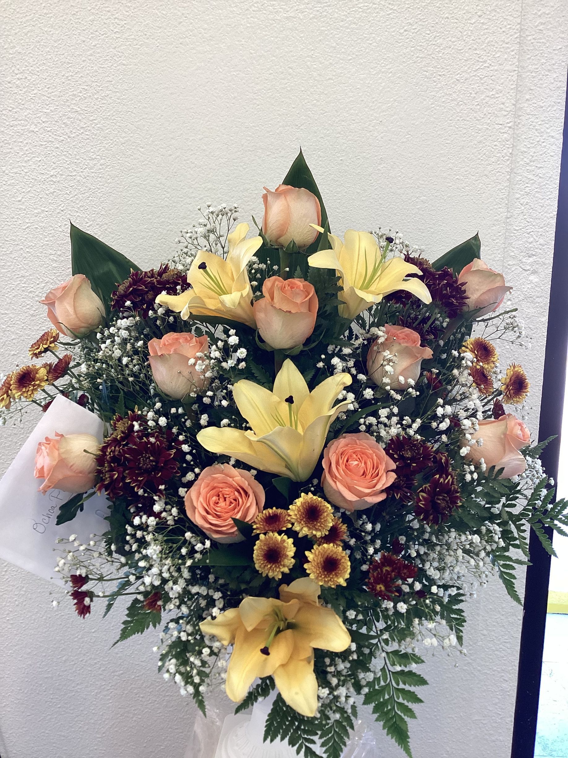 A floral arrangement featuring soft peach roses, yellow lilies, dark red flowers, and baby's breath against a white wall.