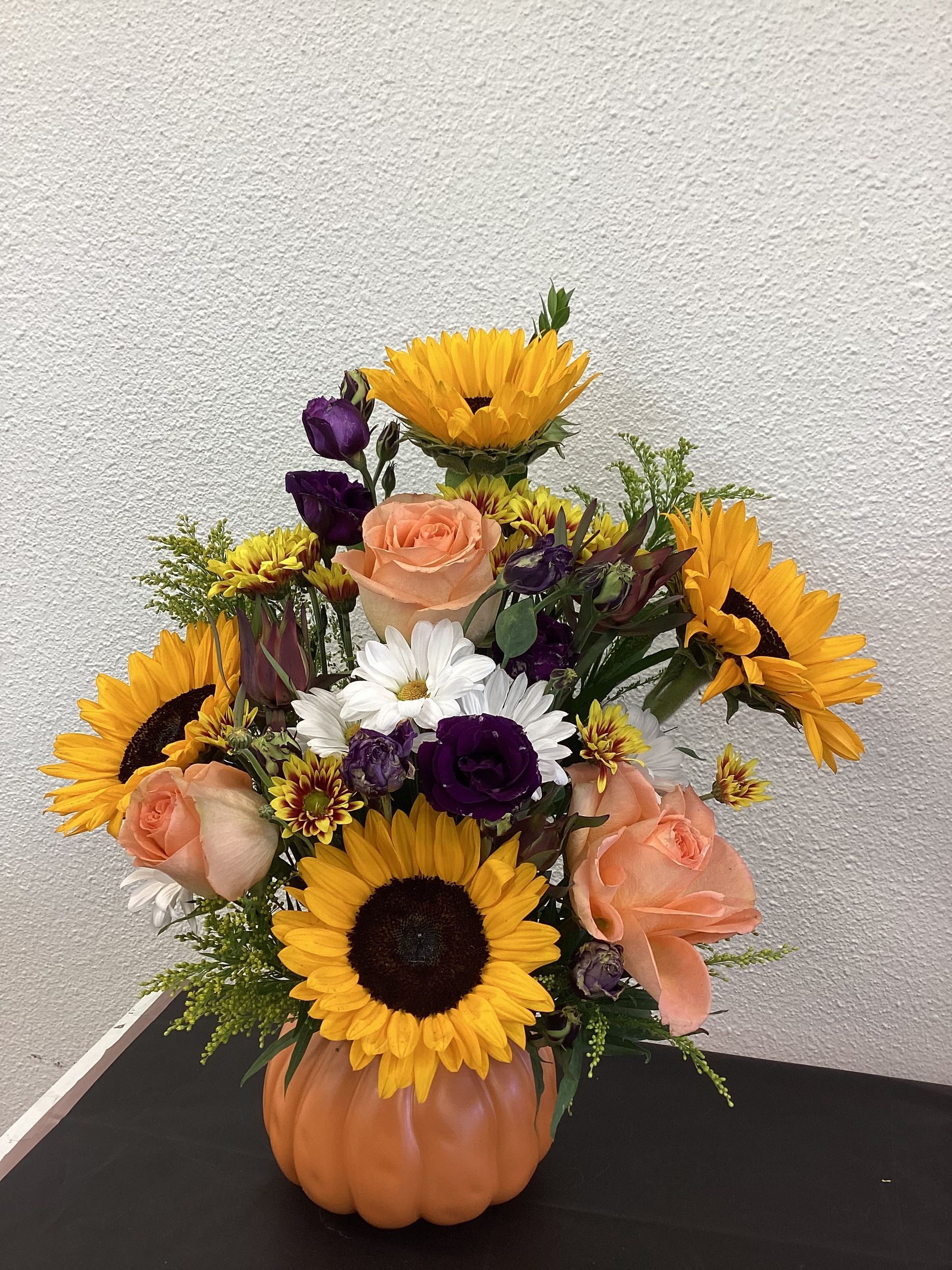 A fall-themed floral arrangement with sunflowers, peach roses, and purple flowers in an orange pumpkin-shaped vase.