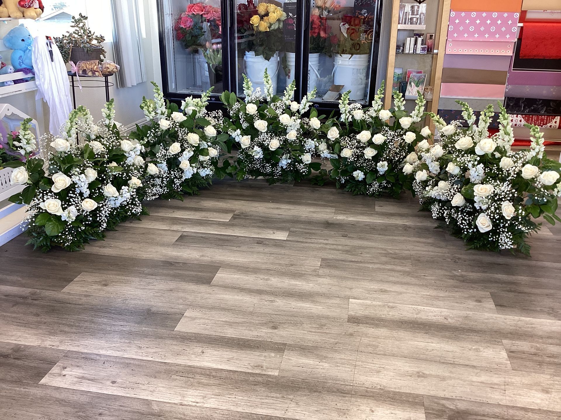 A long, curved floral arrangement of white roses and greenery displayed on a wooden floor in a flower shop.