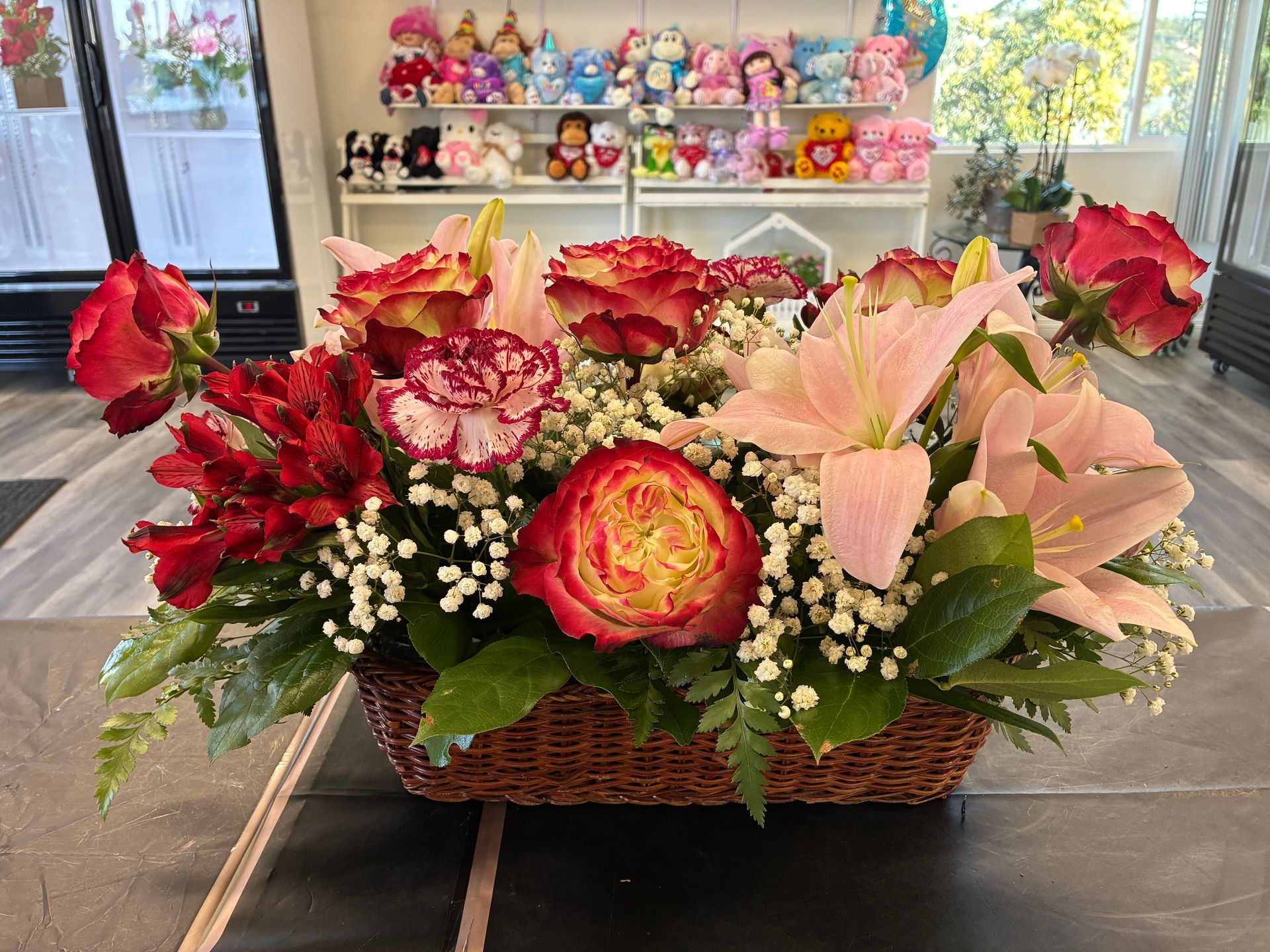 A basket of mixed flowers, including red and yellow roses, pink lilies, and baby’s breath, set on a table.