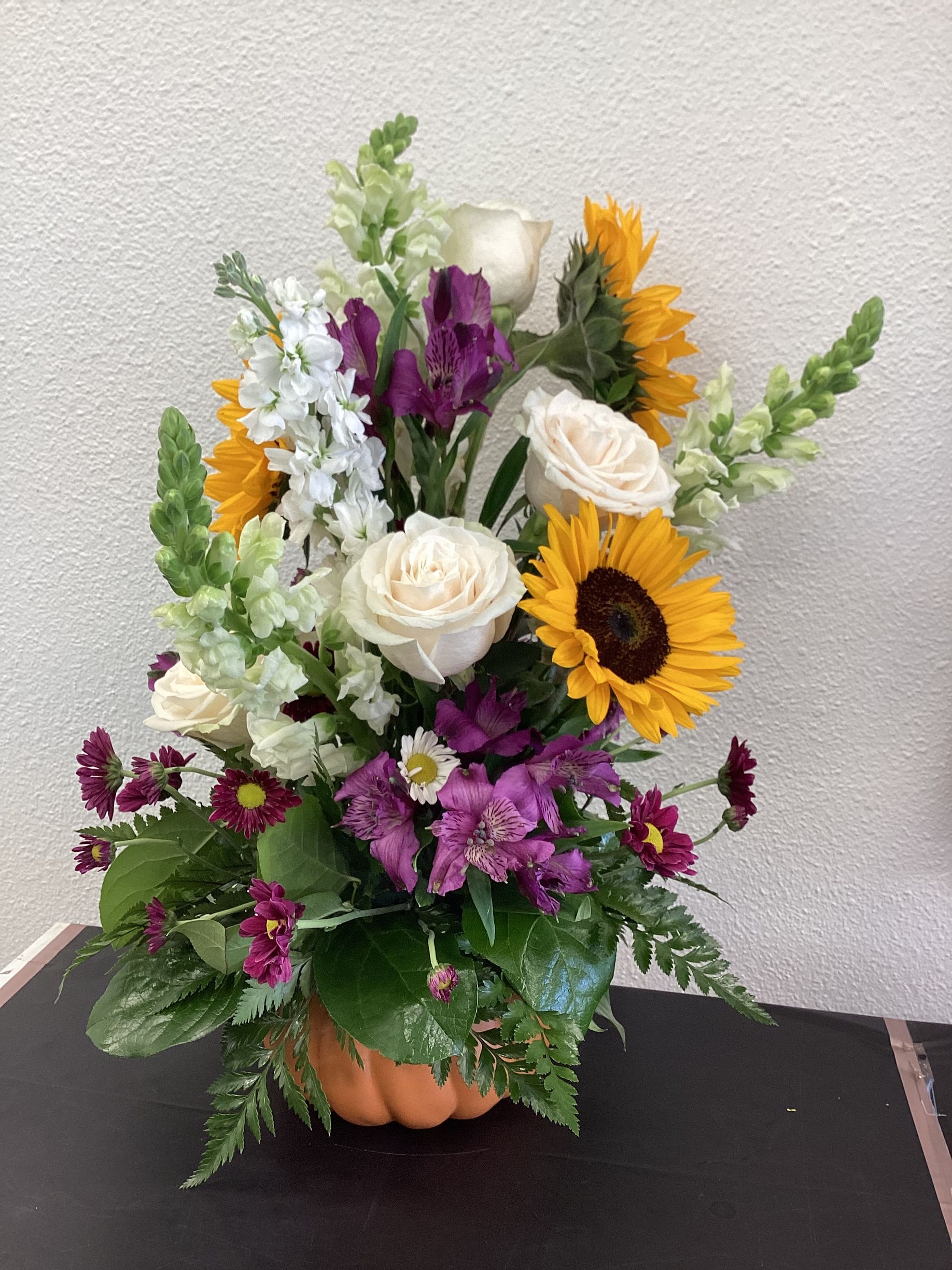 A vibrant floral arrangement in a small pumpkin-shaped vase featuring yellow sunflowers, white roses, and purple flowers.