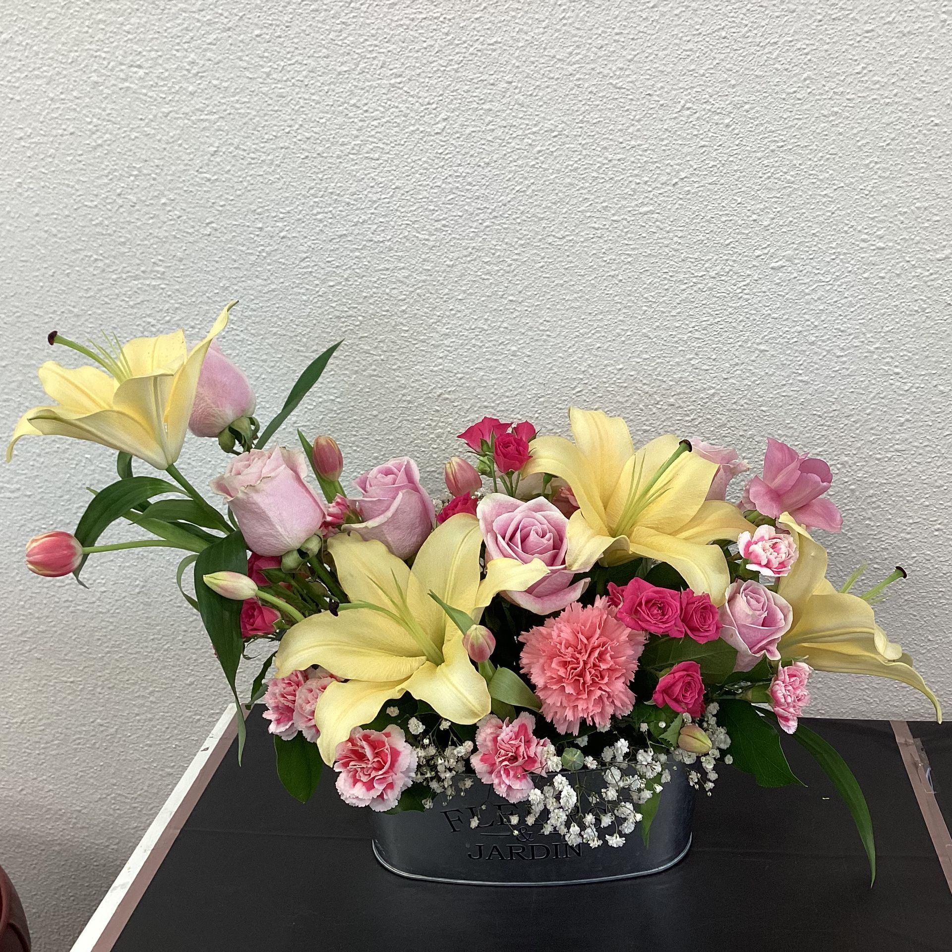 A floral arrangement with yellow lilies, pink roses, and carnations in a small metal bucket against a gray wall.