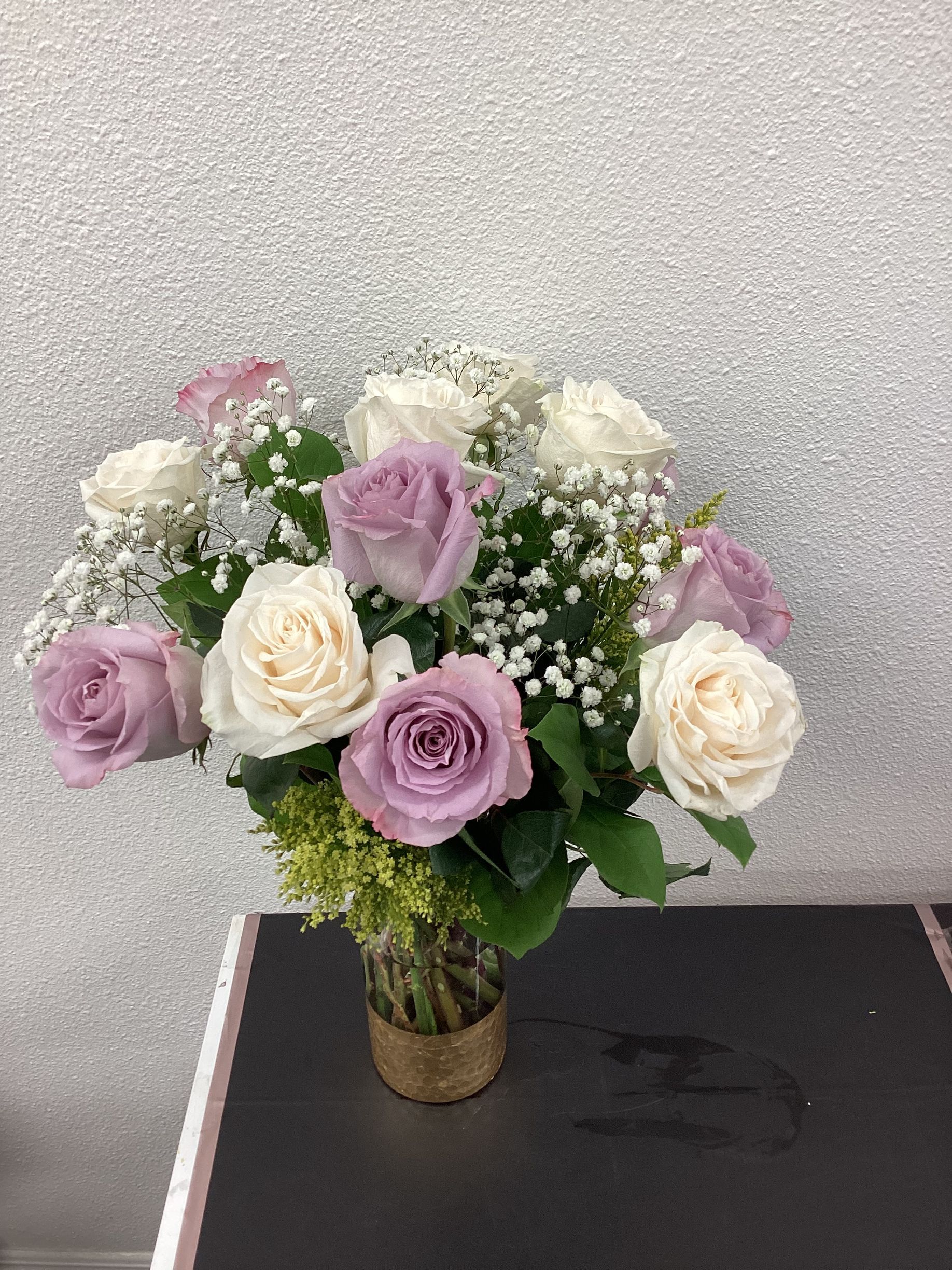 A bouquet of lavender and white roses with baby’s breath in a gold-textured vase, set on a dark surface.