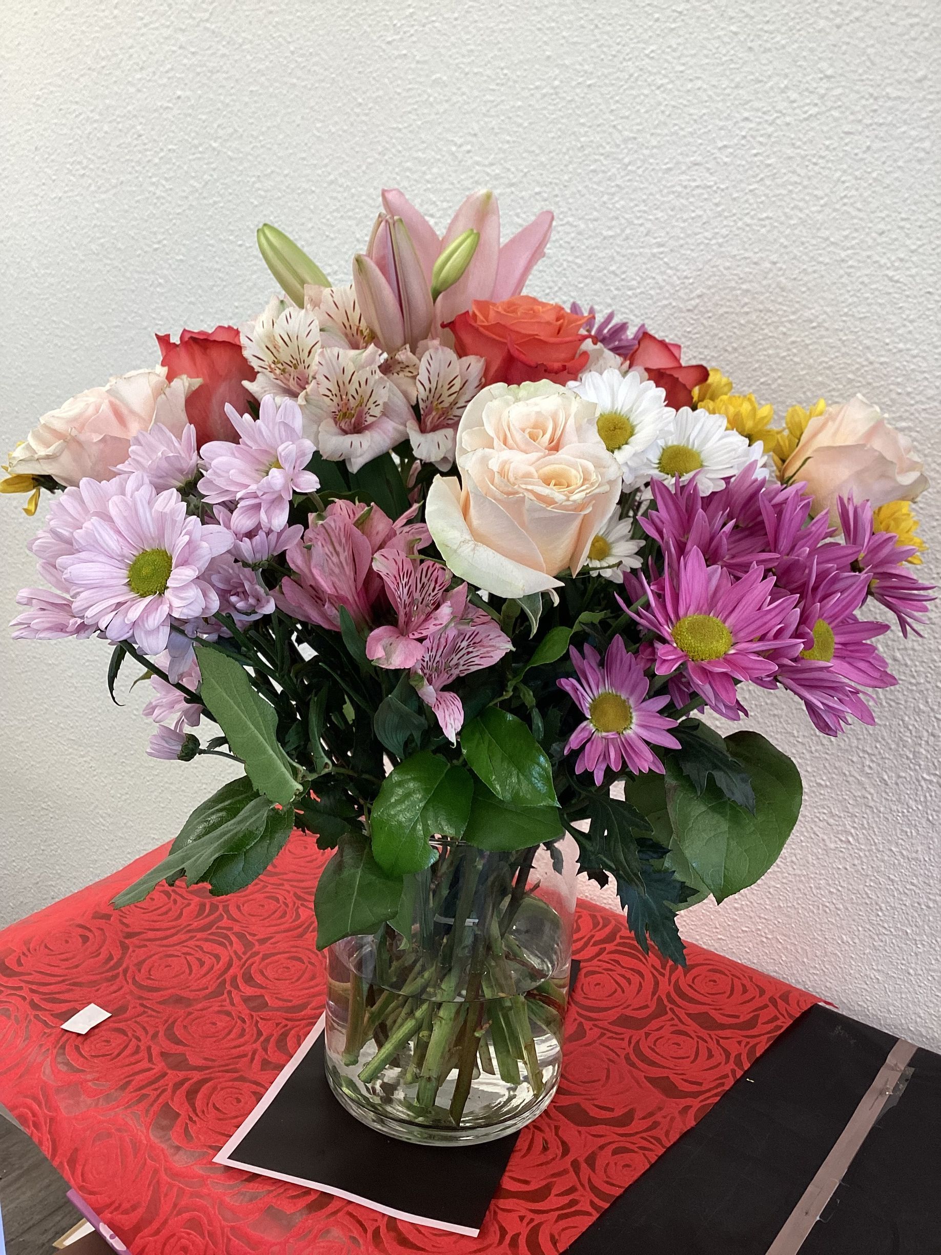 A vibrant floral arrangement in a glass vase, featuring pink lilies, peach roses, and purple and white daisies.