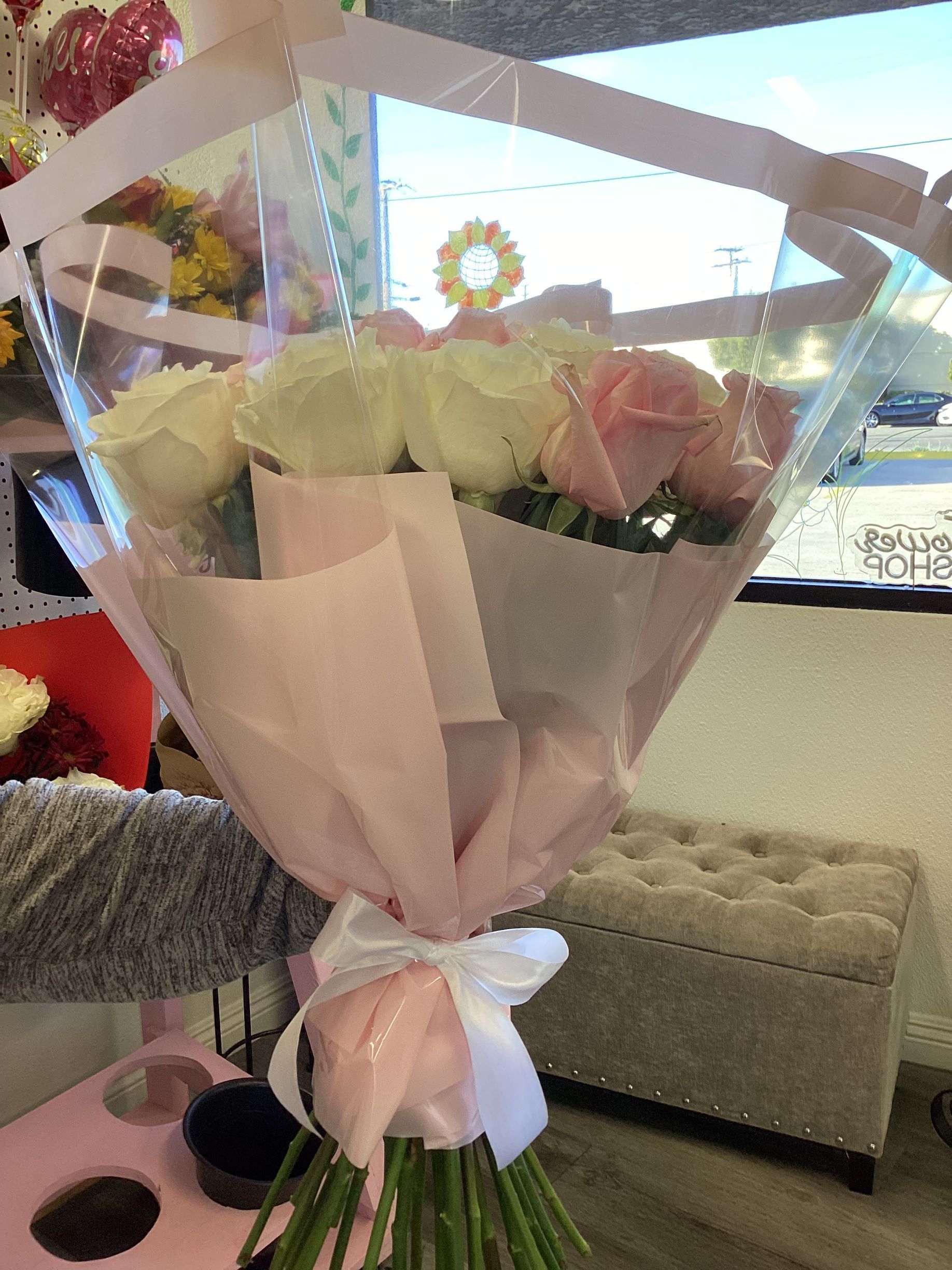 A bouquet of white and pink roses wrapped in clear and pink paper, tied with a white ribbon in a floral shop.