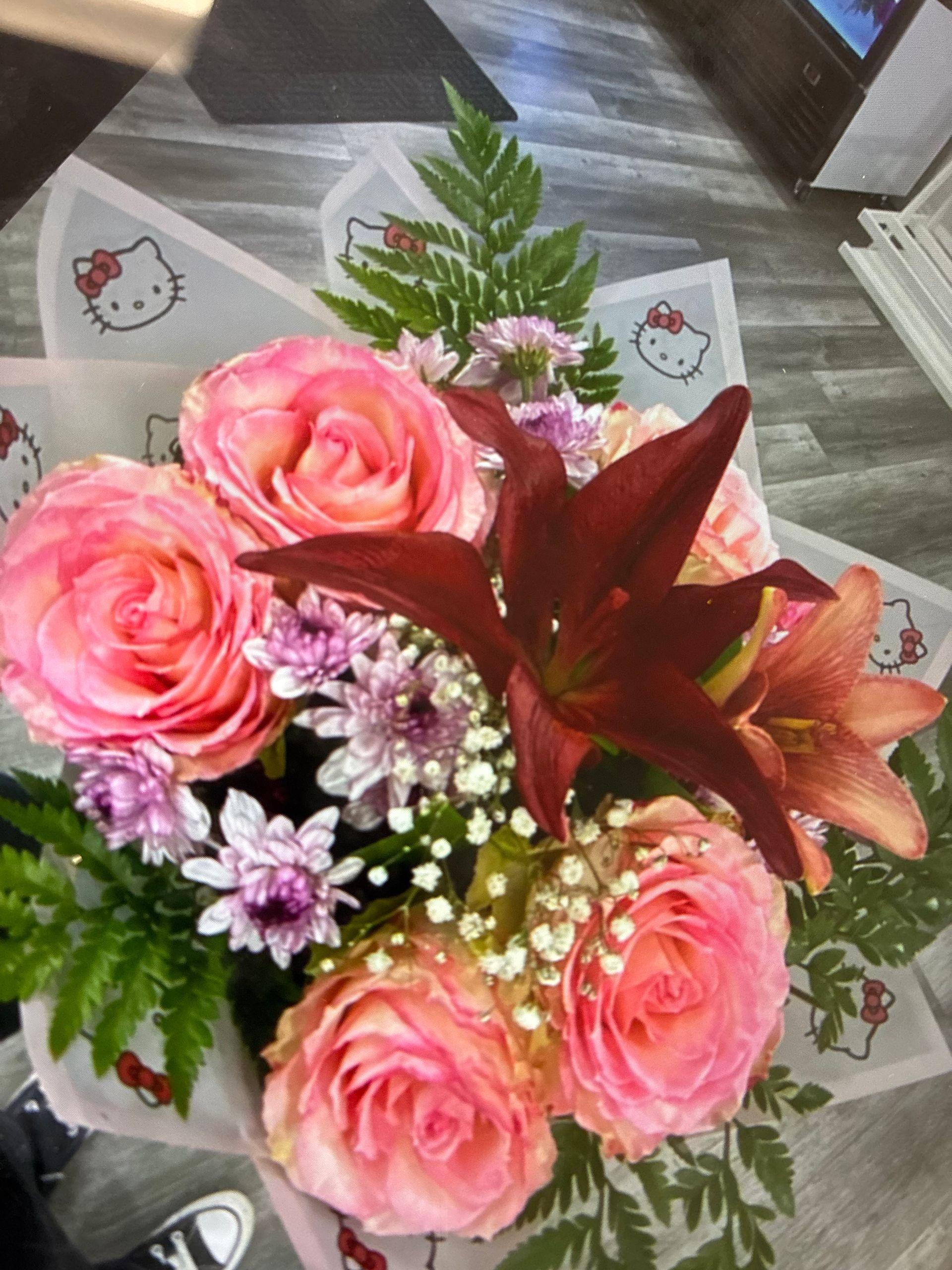 A bouquet of pink roses, a dark red lily, and purple chrysanthemums wrapped in Hello Kitty patterned paper.