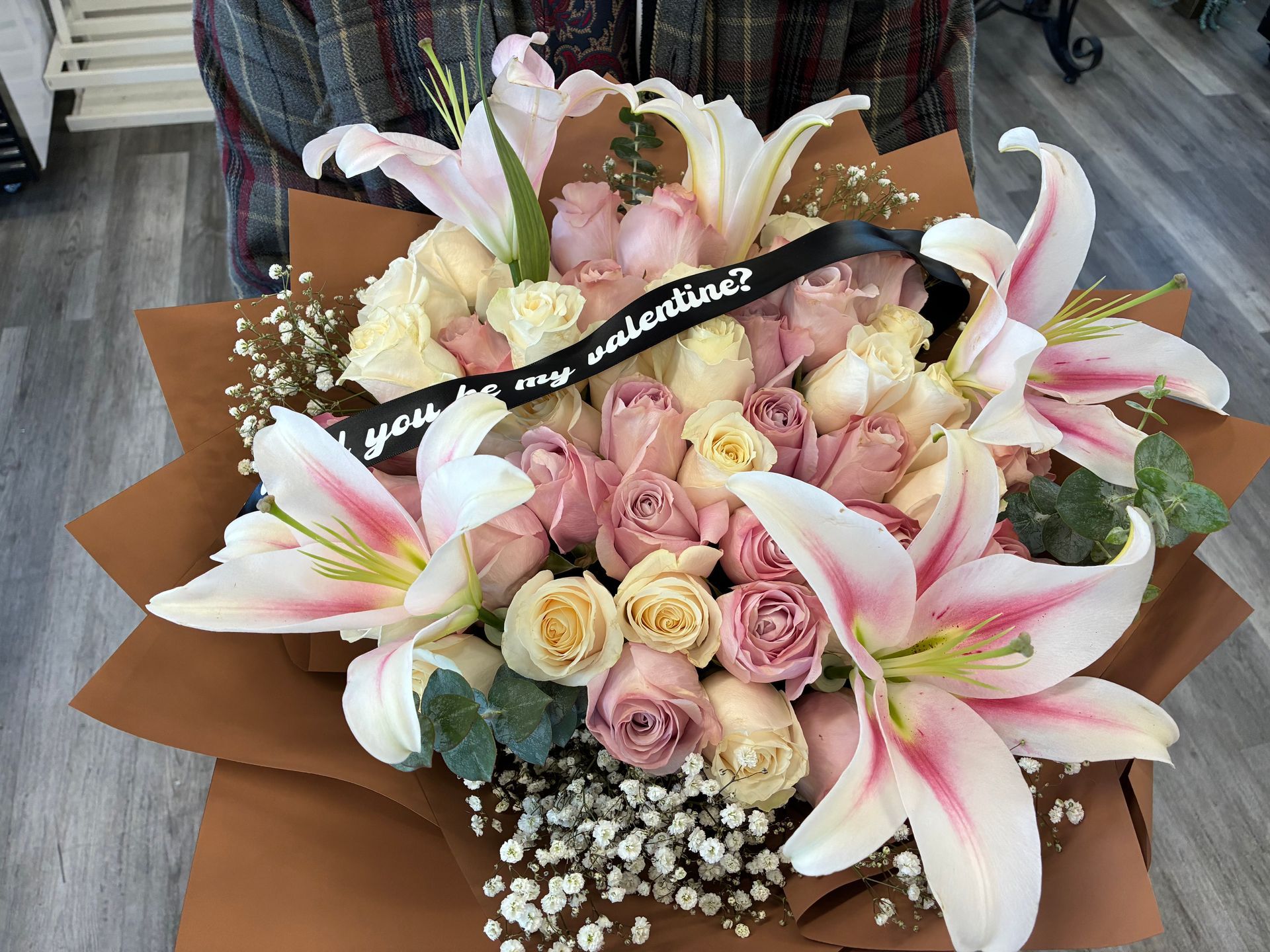 A bouquet of pink and white lilies and roses in brown paper, with a black ribbon reading 