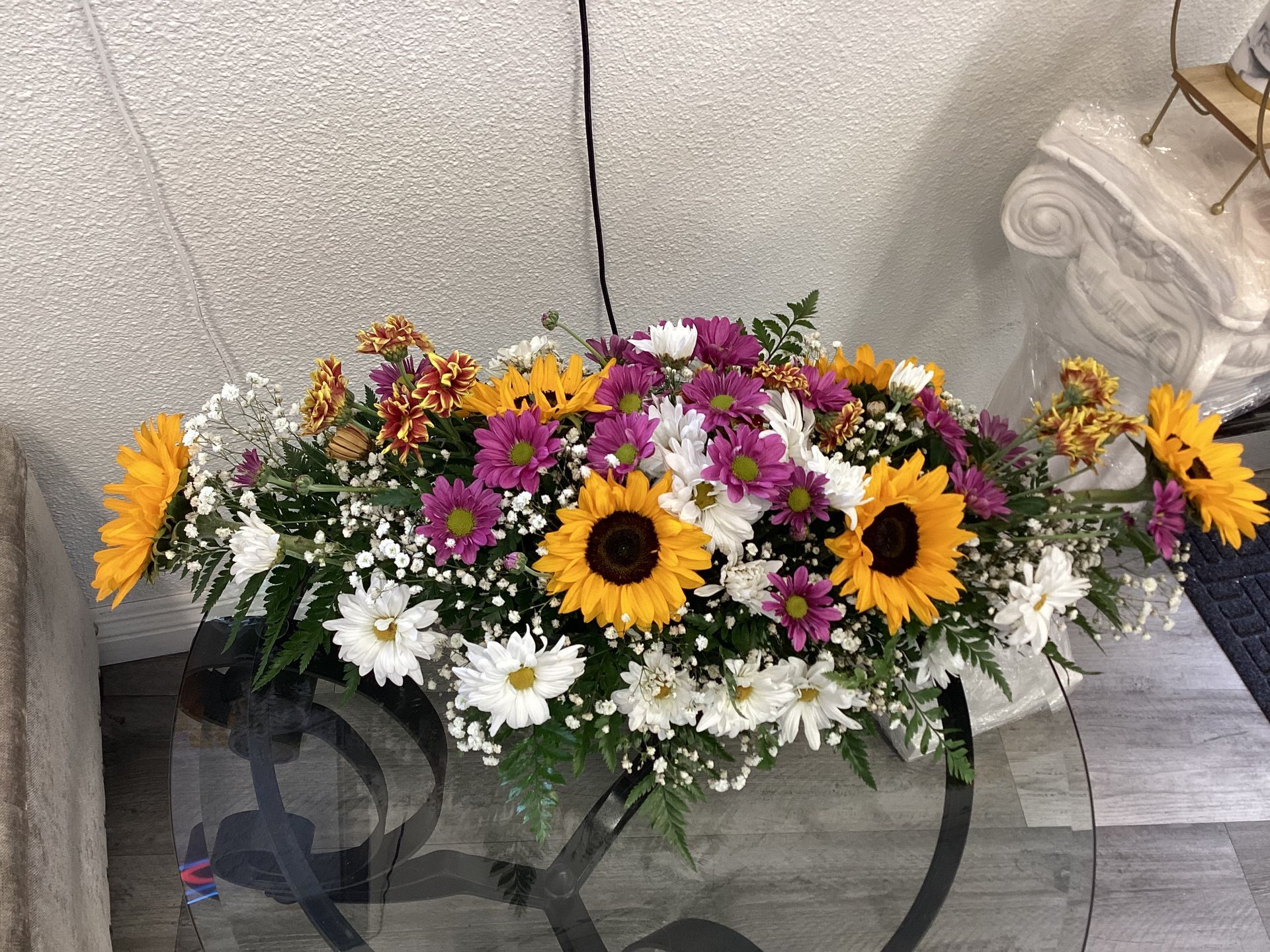 A floral arrangement featuring sunflowers, white daisies, and purple flowers on a glass table against a textured wall.