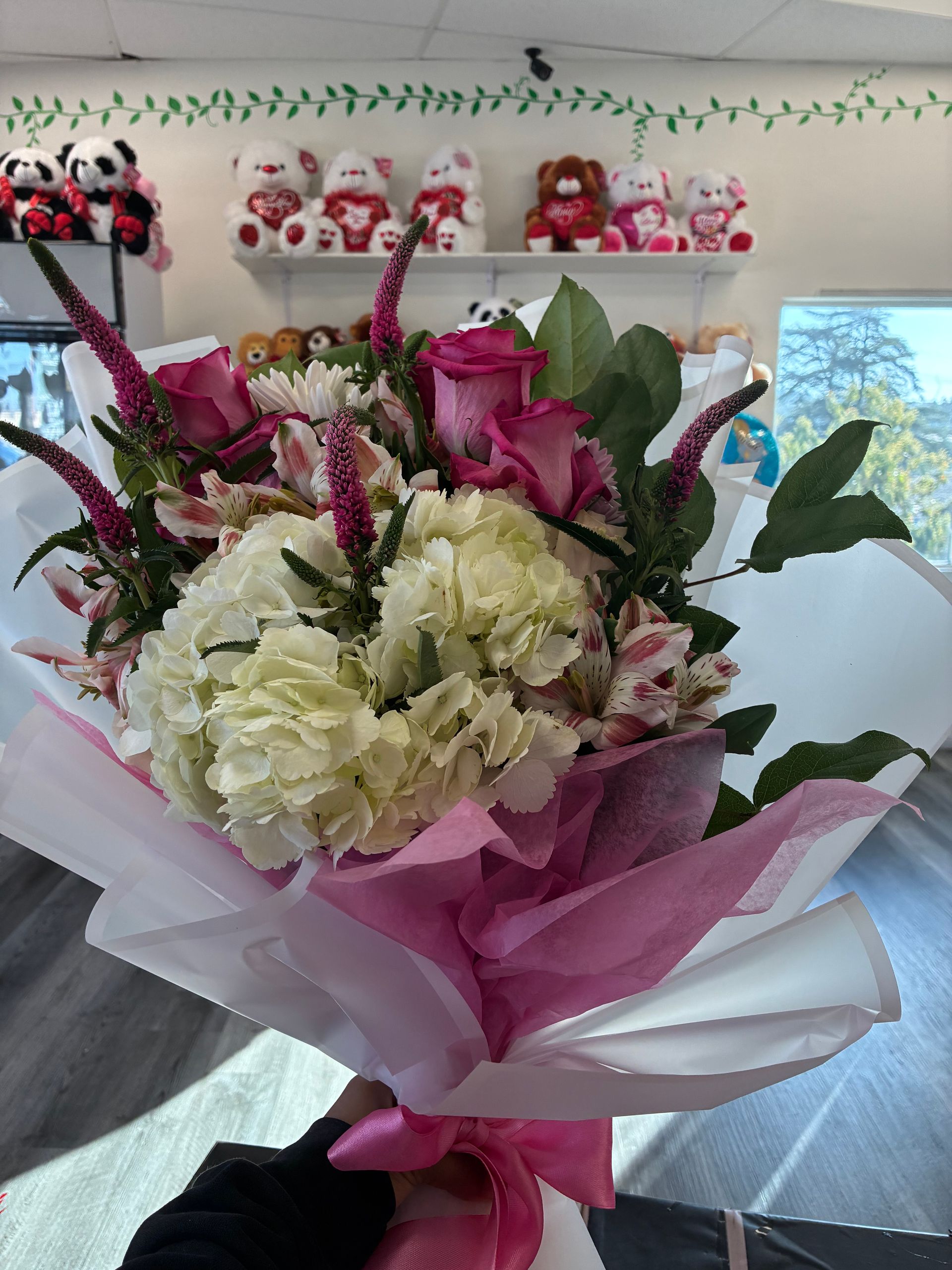 A vibrant bouquet of pink roses, white hydrangeas, and purple accents wrapped in white and pink paper, held in a shop.