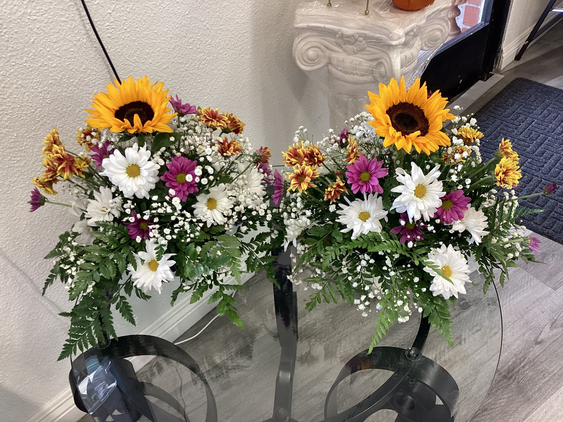 Two symmetrical floral arrangements with sunflowers, white daisies, purple asters, and baby's breath on a glass table.