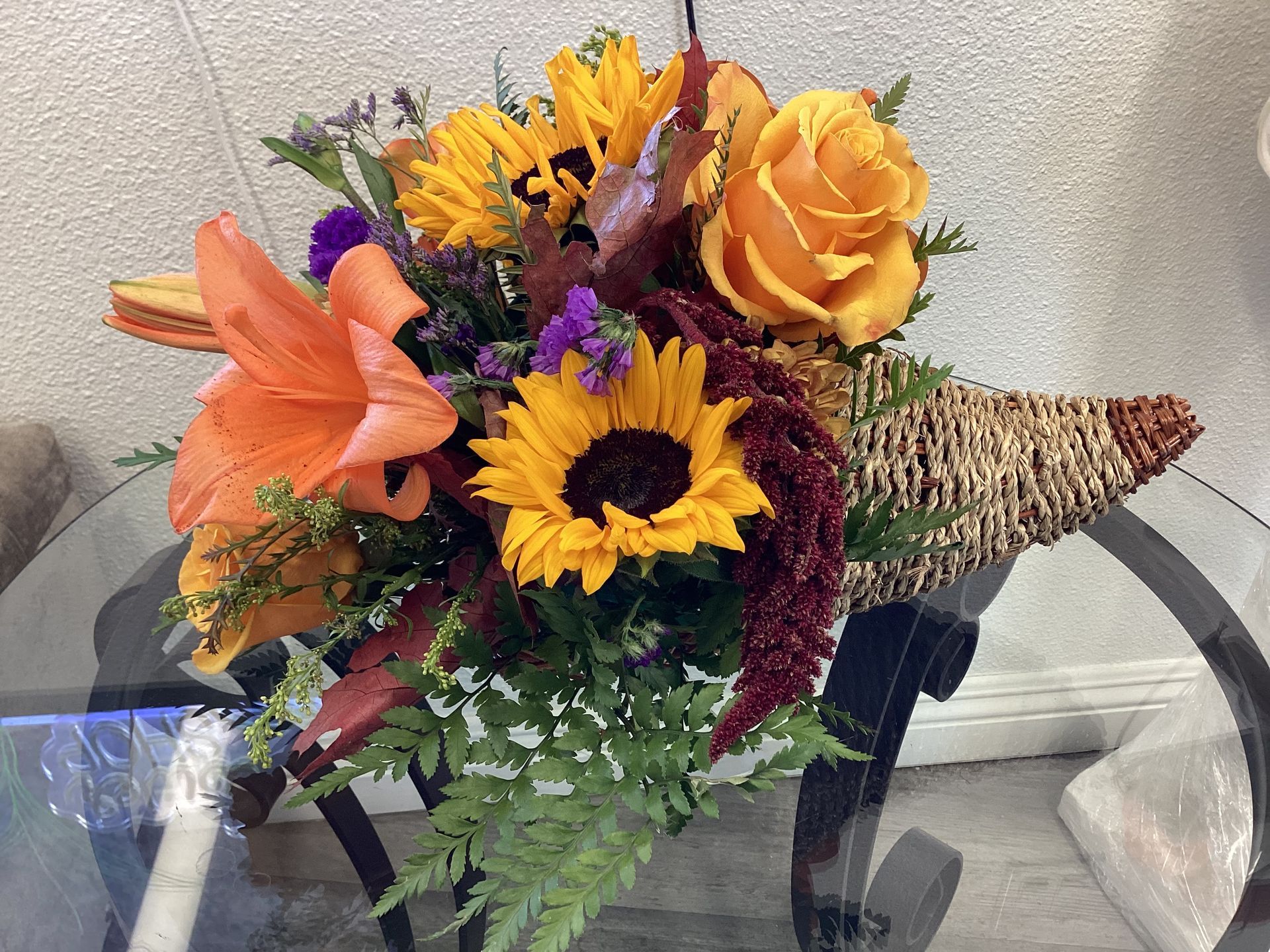 A fall-themed cornucopia arrangement with orange lilies, sunflowers, yellow roses, and greenery on a glass table.