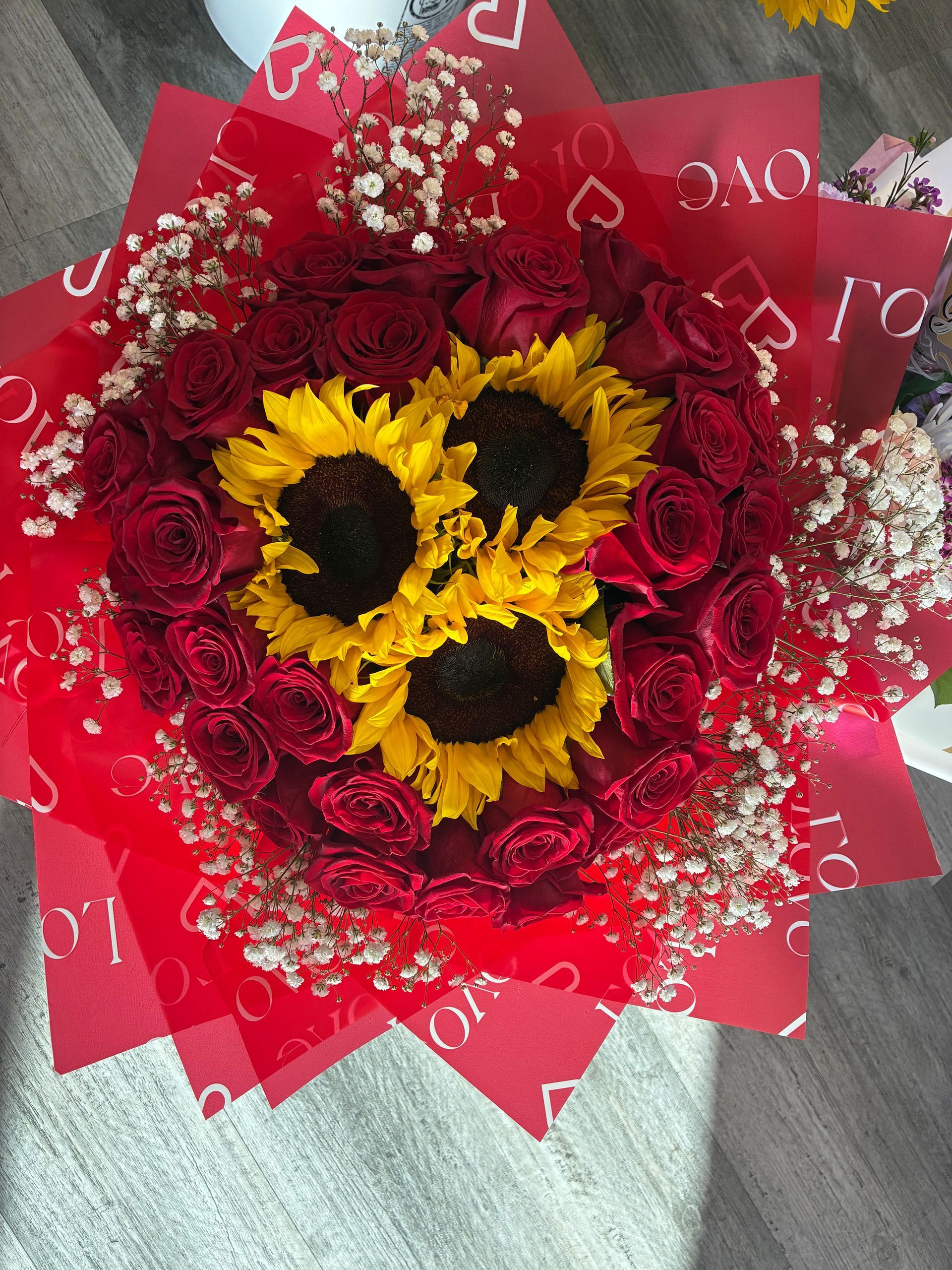 A bouquet featuring three sunflowers surrounded by red roses and baby's breath, wrapped in red 