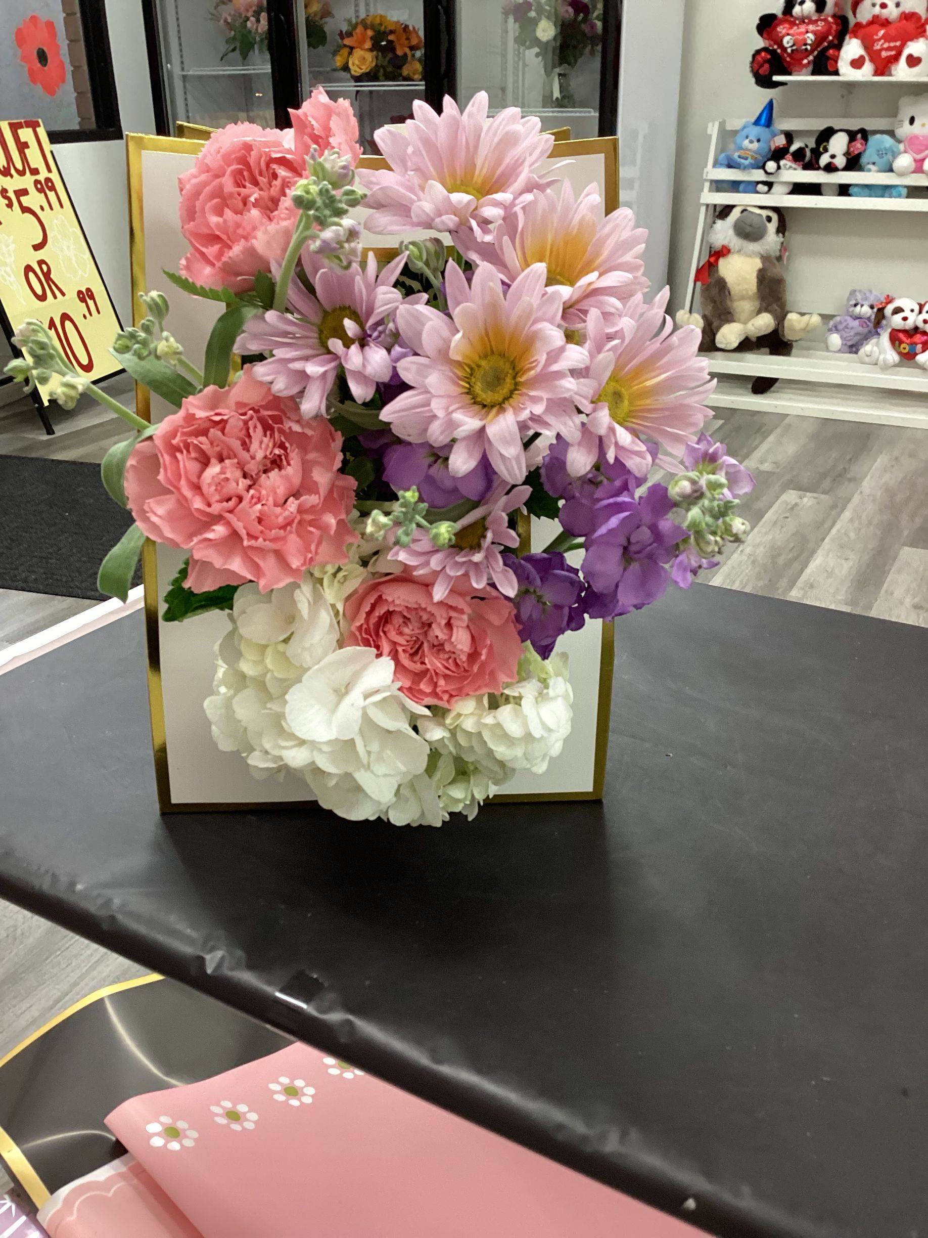 A floral arrangement in a white and gold box, featuring pink carnations, light purple daisies, and white hydrangeas.