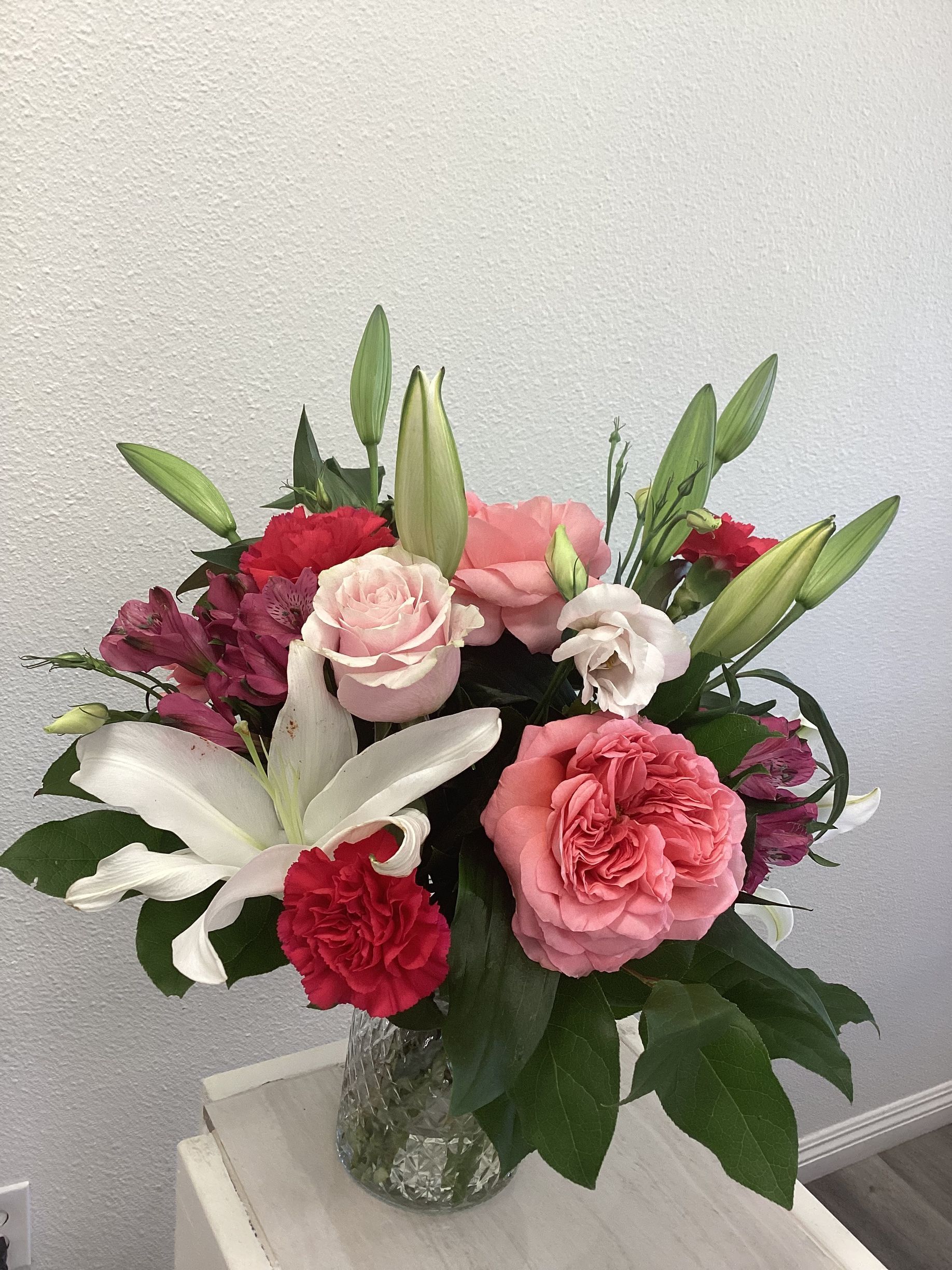 A colorful floral arrangement in a glass vase featuring white lilies, pink and red roses, and carnations against a wall.