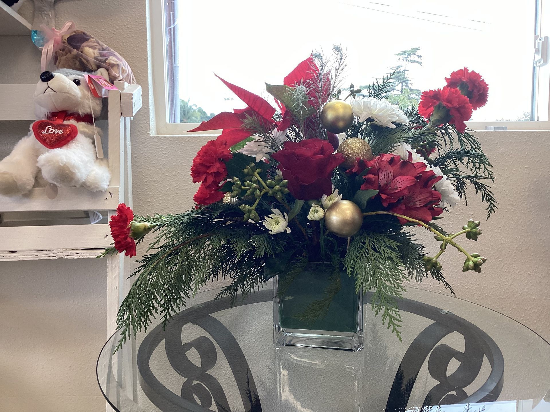 A floral arrangement with red roses, carnations, and greenery in a glass vase, sitting on a table near a white teddy bear.