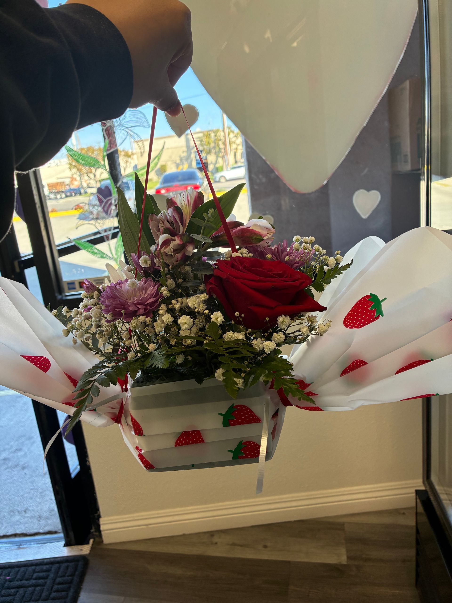 A hand holds a small floral arrangement with a single red rose, baby's breath, and greenery wrapped in strawberry fabric.