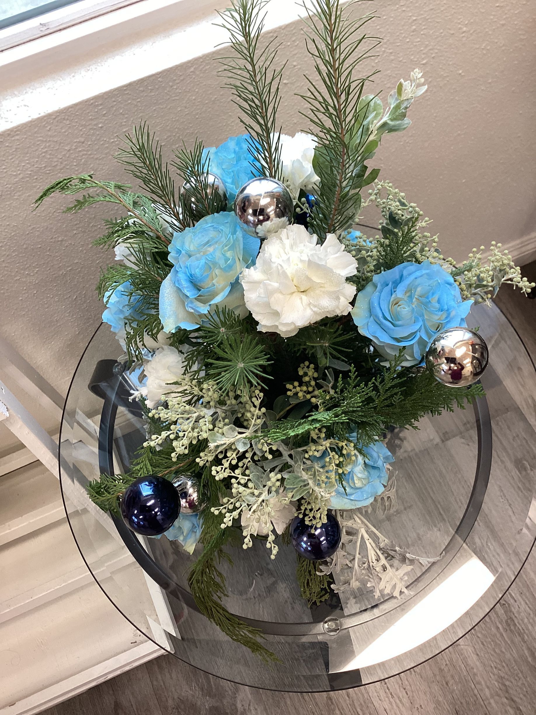 A floral arrangement with blue and white roses, greenery, and small silver and blue ornaments on a round glass table.