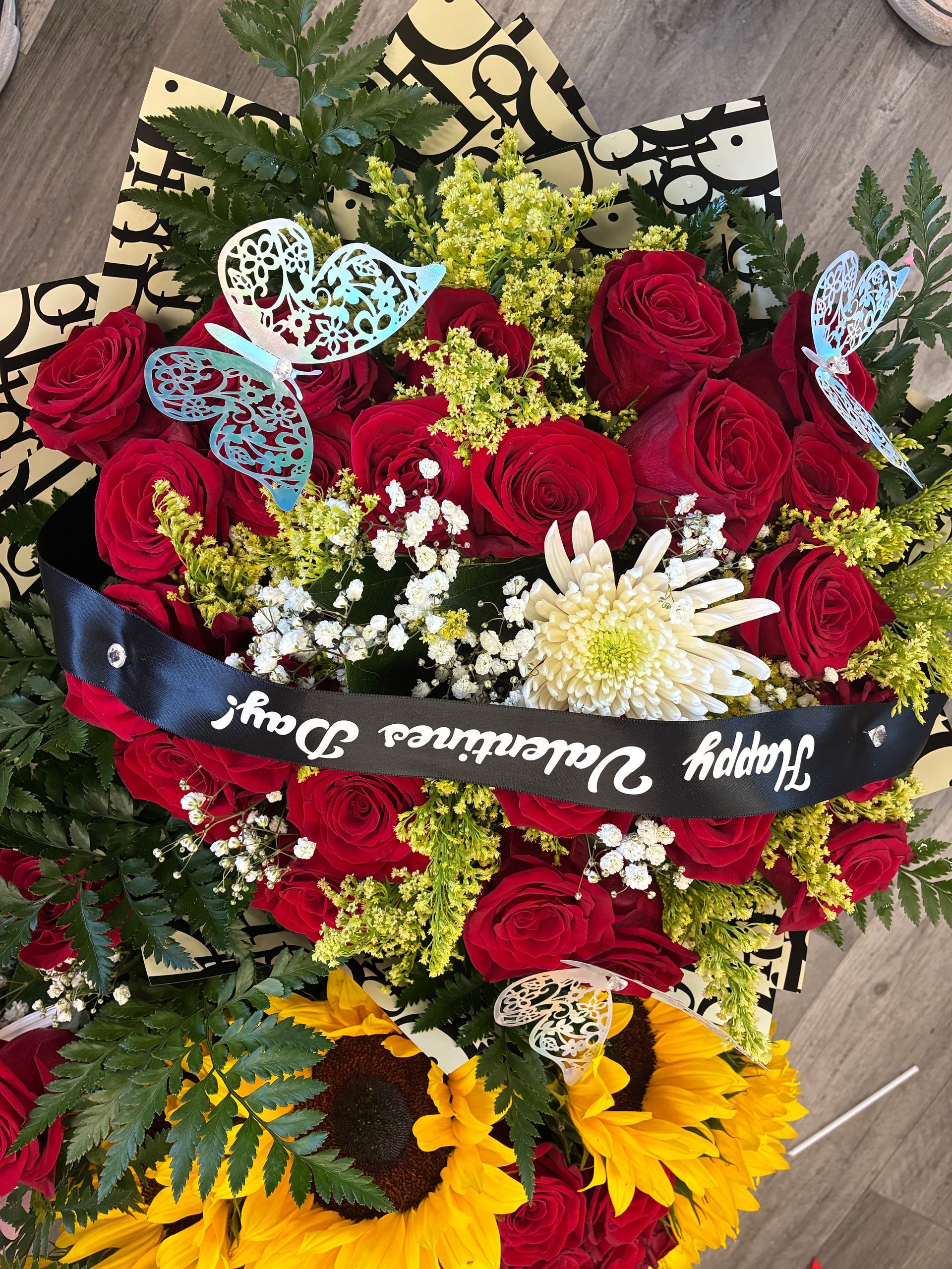 A bouquet of red roses, sunflowers, and baby's breath, decorated with two blue butterfly ornaments and a Valentine's ribbon.