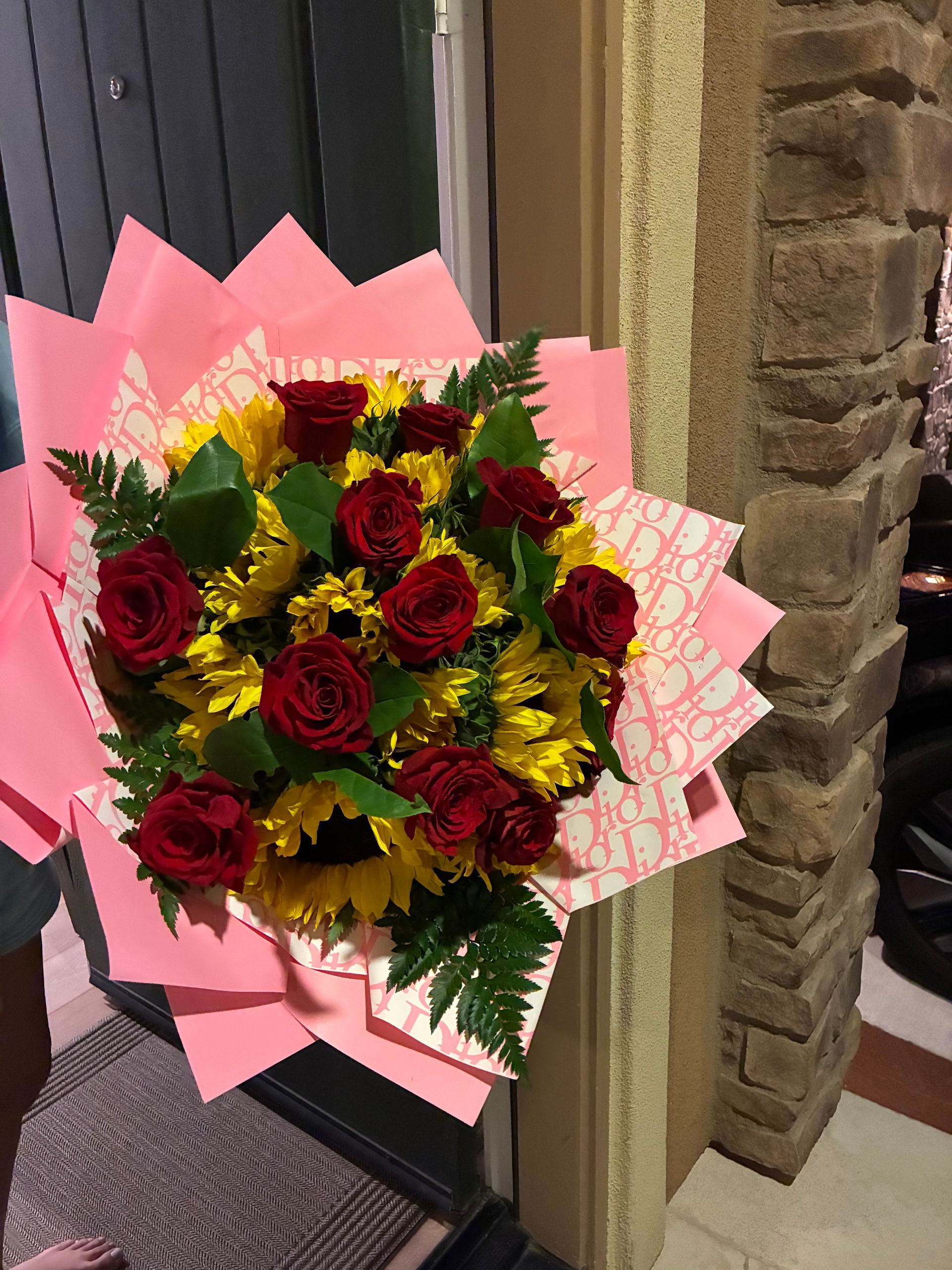 A vibrant bouquet of red roses and yellow sunflowers wrapped in pink, patterned paper, held near a doorway.