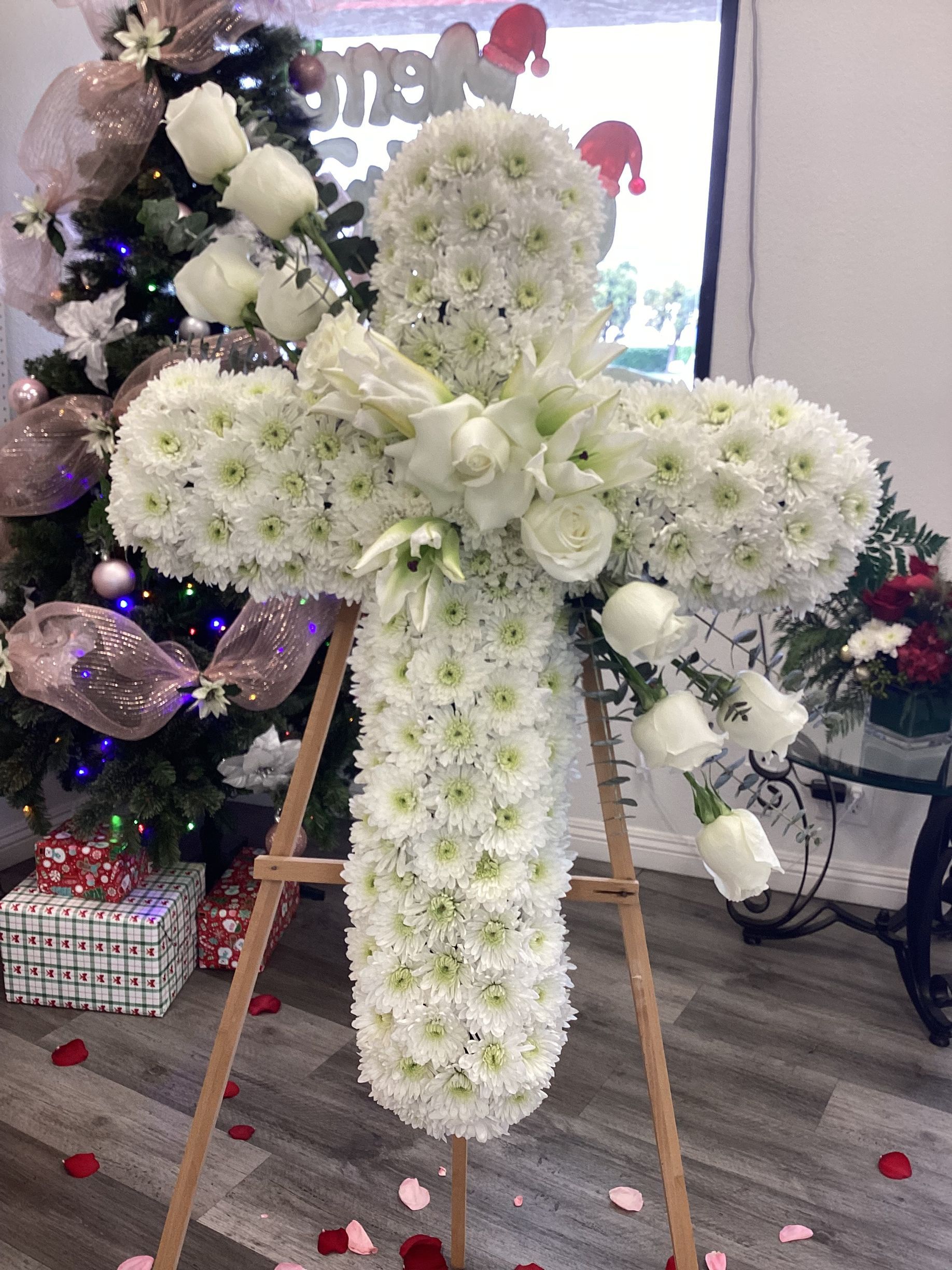A large white floral cross arrangement on a wooden easel, placed in a room decorated with a holiday tree and wrapped gifts.