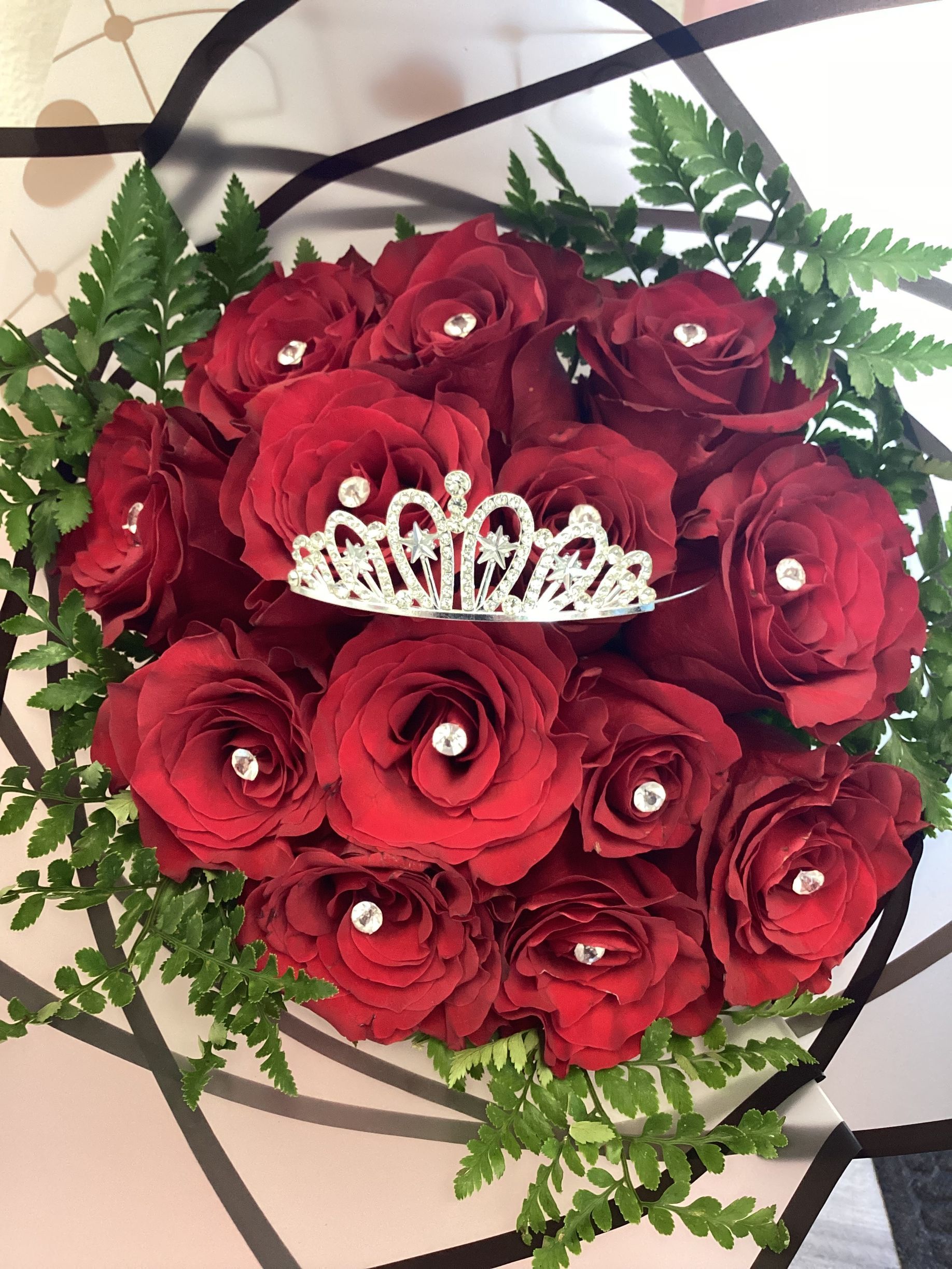 A bouquet of deep red roses accented with sparkling pins, topped with a decorative silver tiara, against a geometric backdrop.