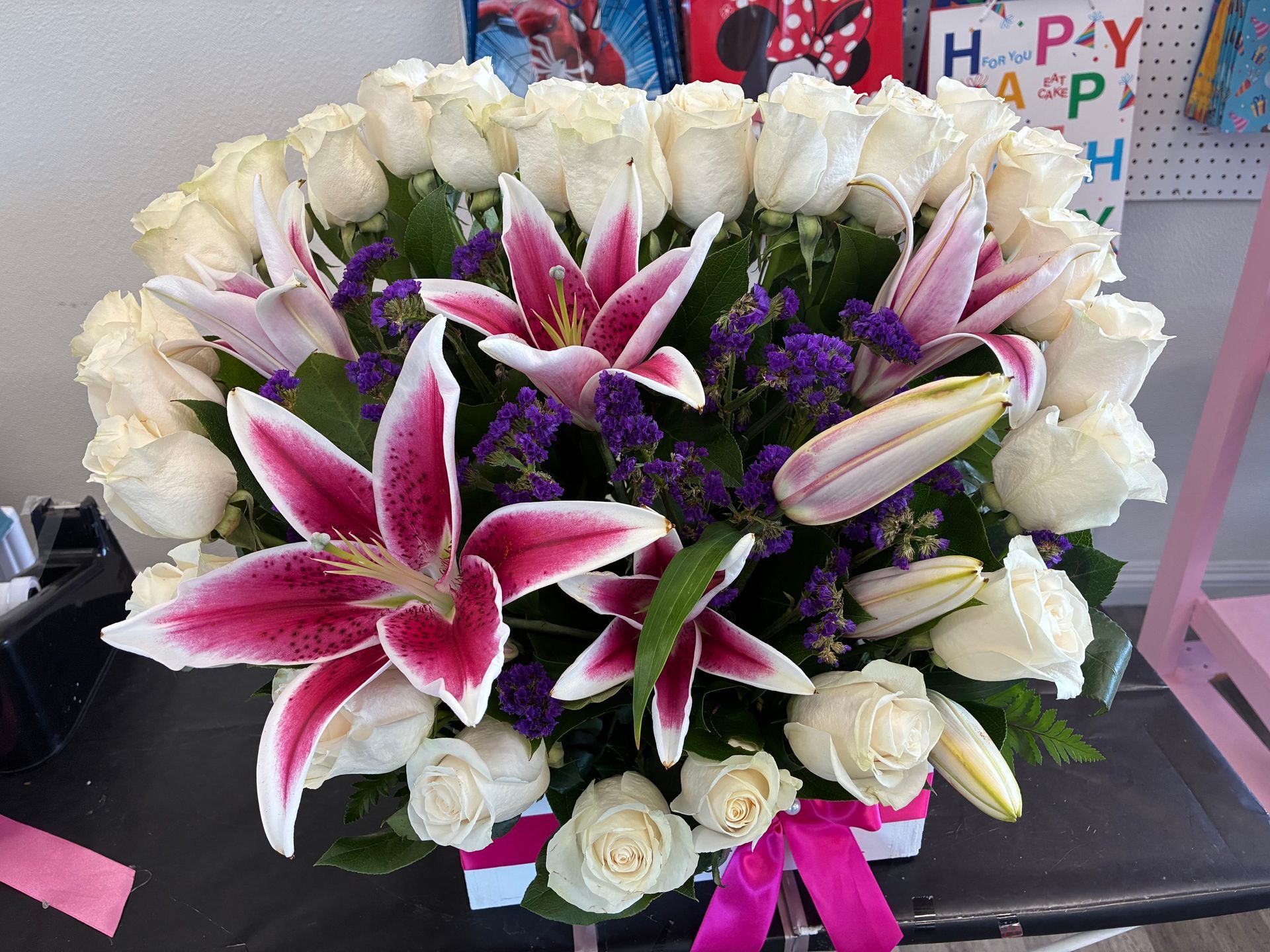 A large, lush bouquet of white roses and pink-spotted lilies accented with purple statice, tied with a bright pink ribbon.