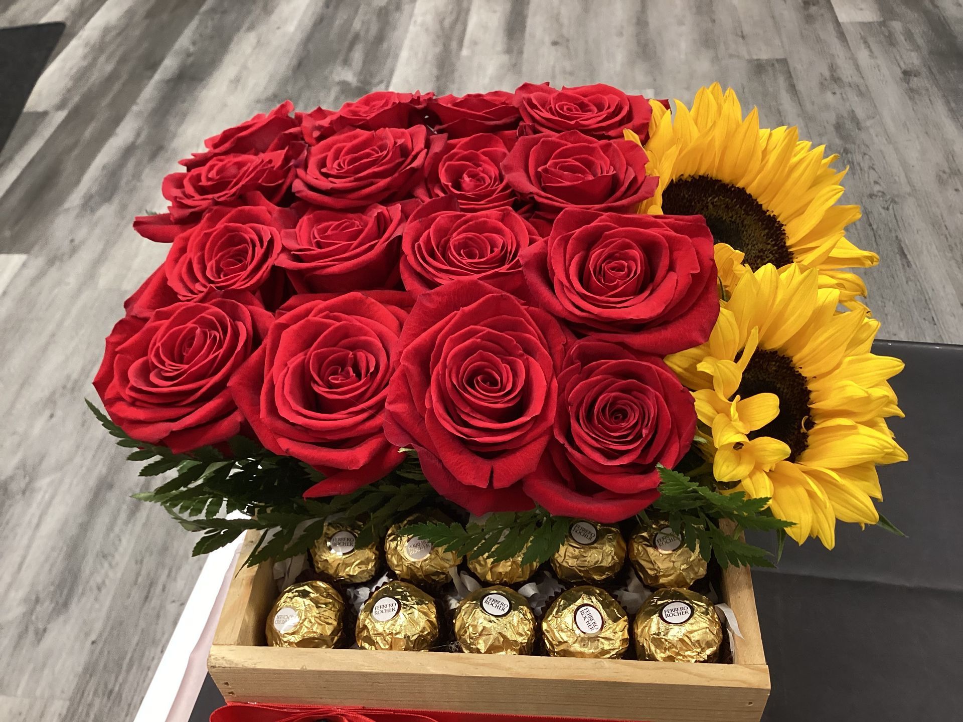 A floral arrangement with red roses and two yellow sunflowers in a wooden box lined with gold-wrapped chocolates.