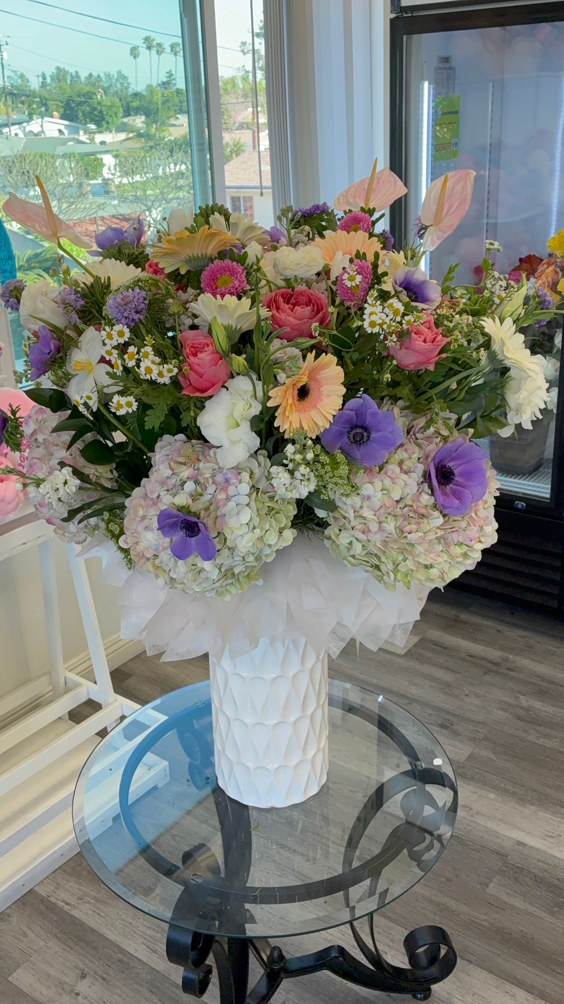 A large, colorful floral arrangement in a white vase, displayed on a glass table in a bright room.
