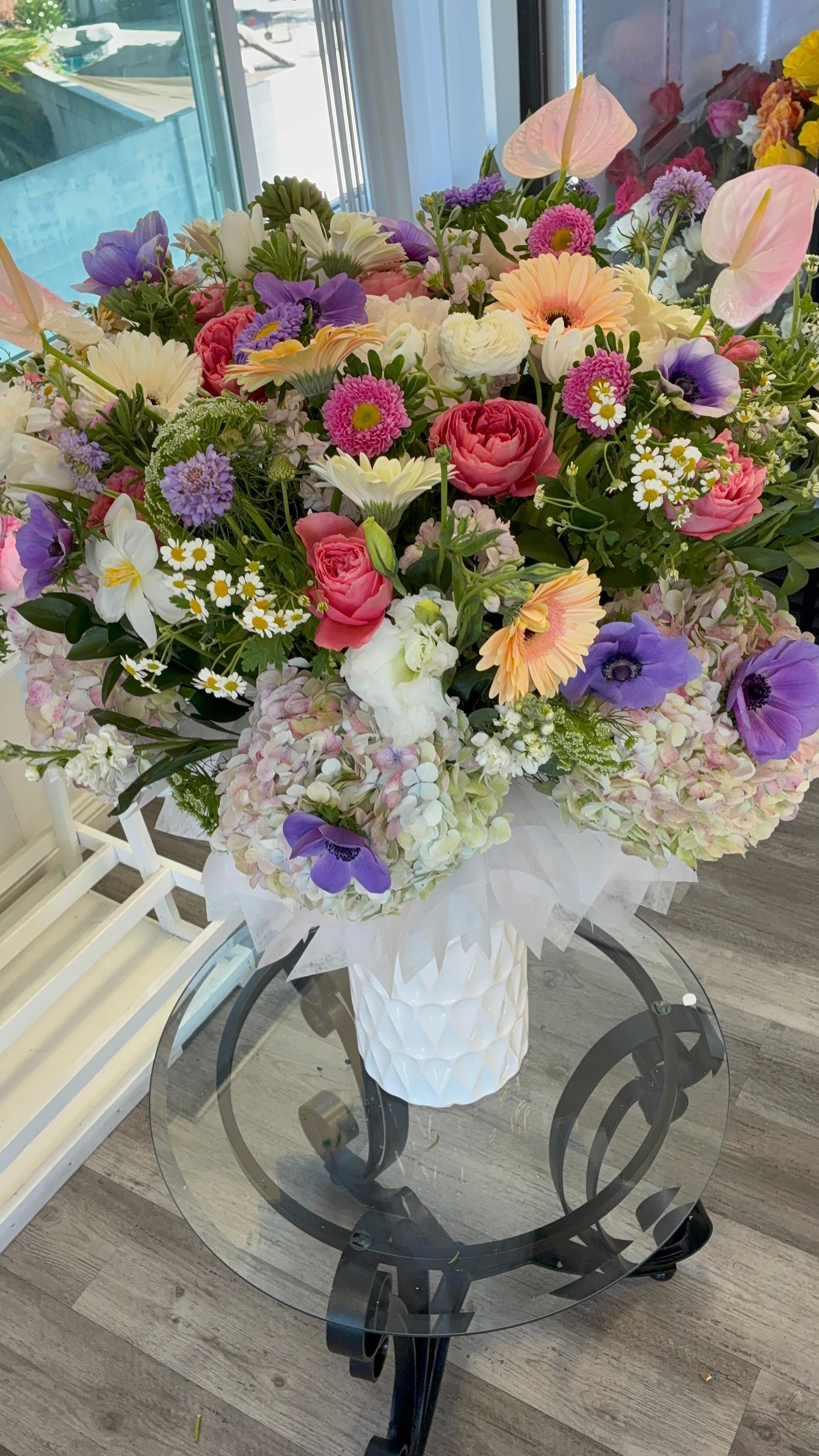 A large, colorful bouquet of assorted flowers in a white textured vase, set on a round glass table with a black metal base.