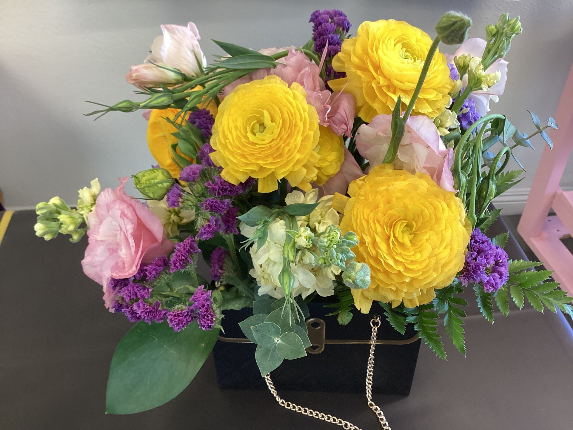 A floral arrangement with yellow ranunculus, pink lisianthus, and purple statice in a black box with a gold chain handle.