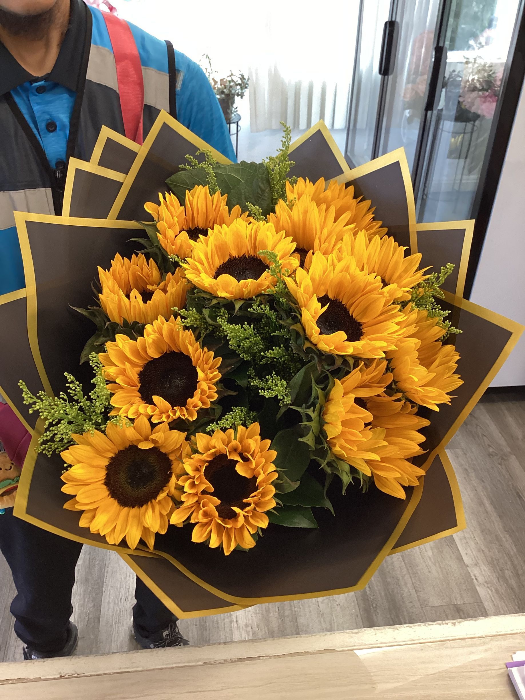 A person holds a large bouquet of sunflowers wrapped in black and gold paper against a store background.