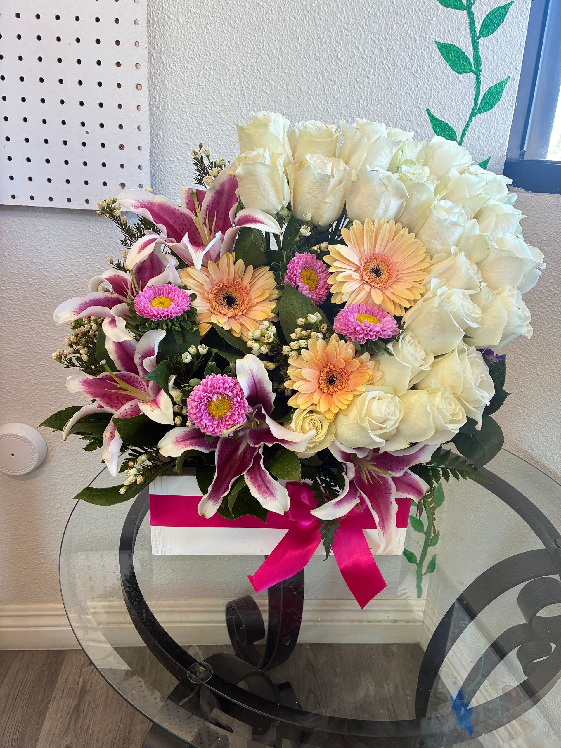 A bouquet of white roses, pink lilies, and orange gerbera daisies in a white box with a bright pink ribbon on a glass table.