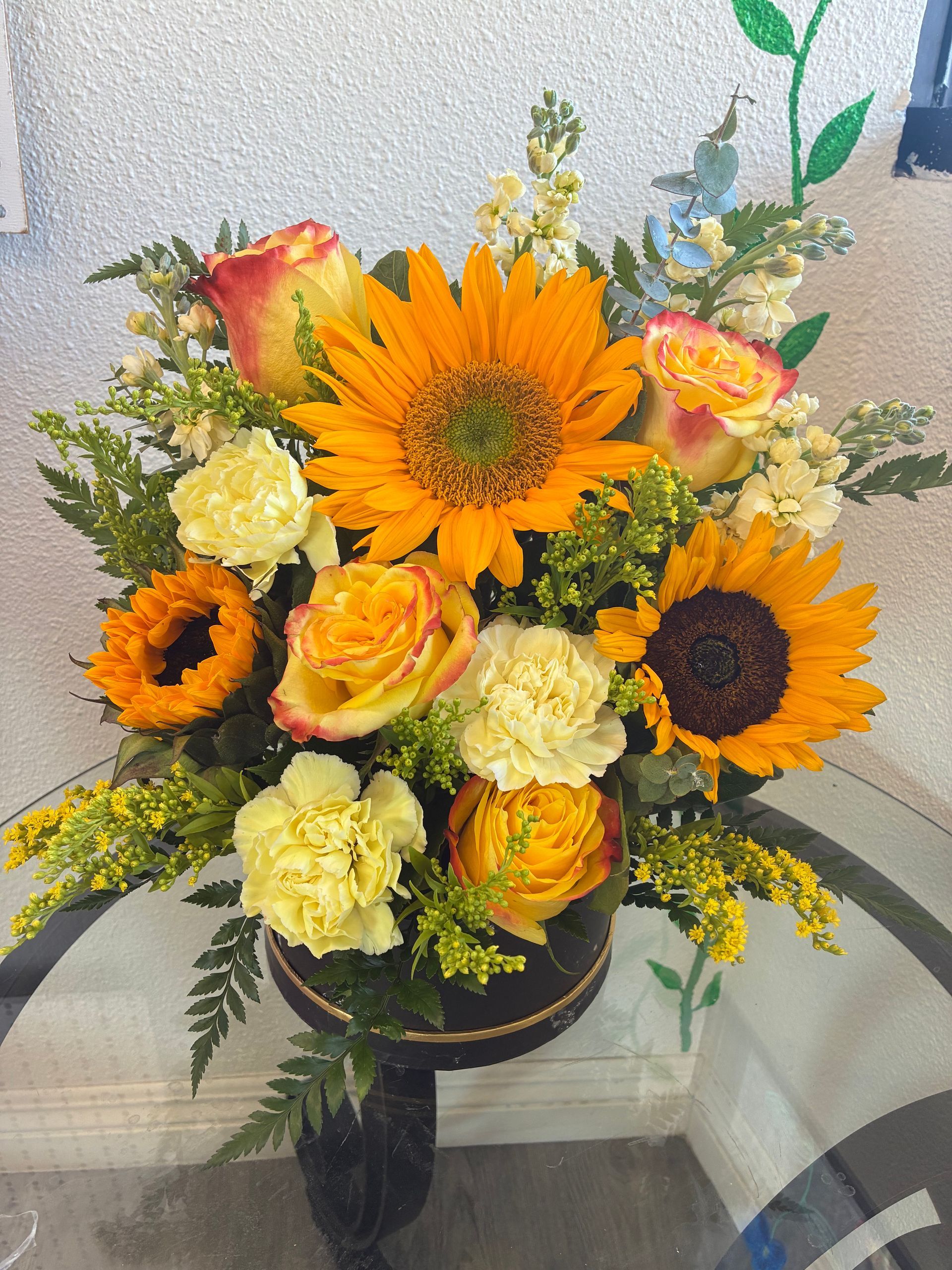 A vibrant floral arrangement featuring yellow sunflowers, orange-edged roses, and carnations in a dark decorative bowl.