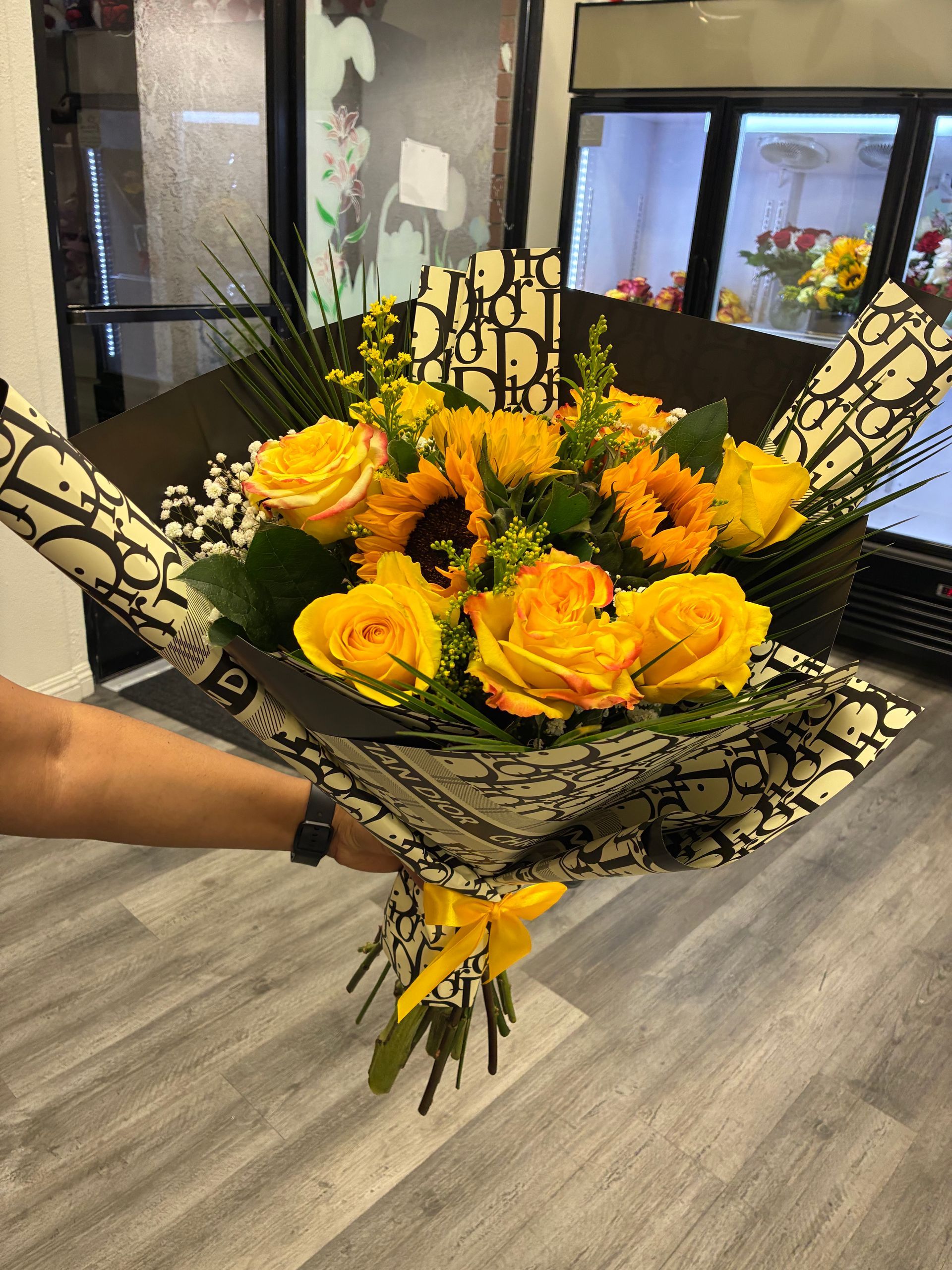 A hand holds a vibrant bouquet of yellow roses and sunflowers, wrapped in patterned black and white paper with a bow.