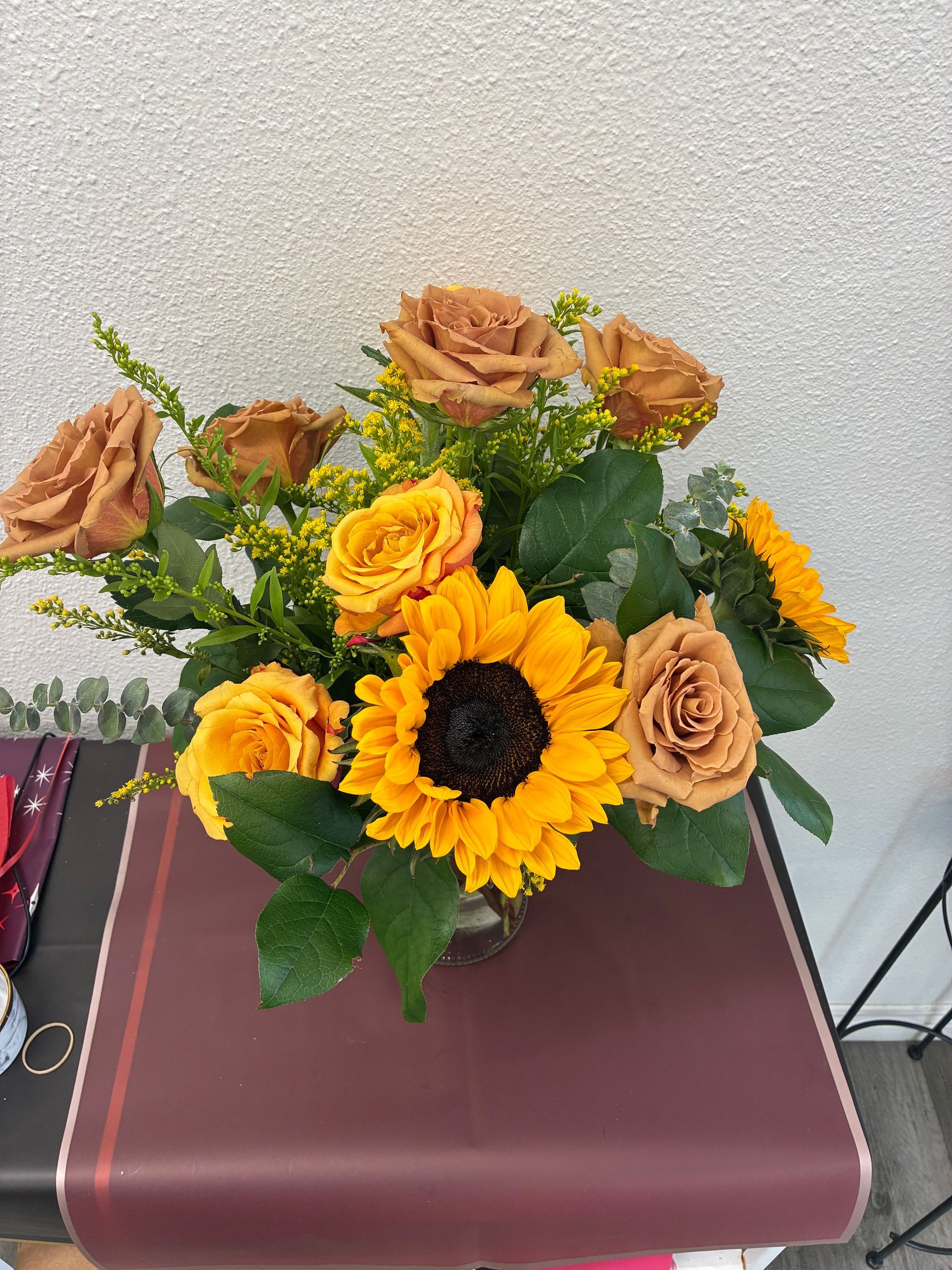A bouquet of tan roses and bright yellow sunflowers mixed with green foliage in a vase on a table.