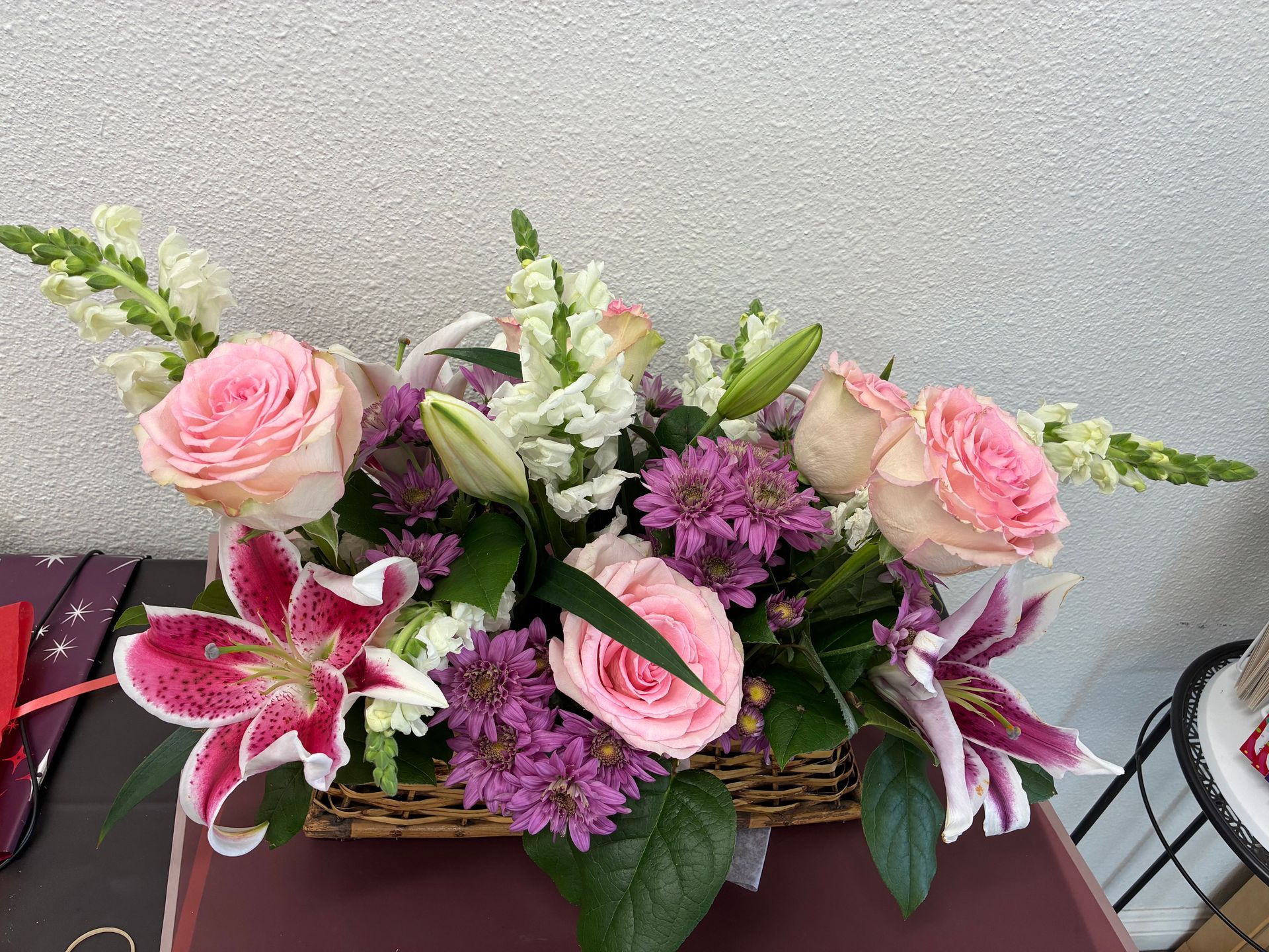 A wicker basket overflowing with a floral arrangement of pink roses, white snapdragons, and purple-spotted stargazer lilies.