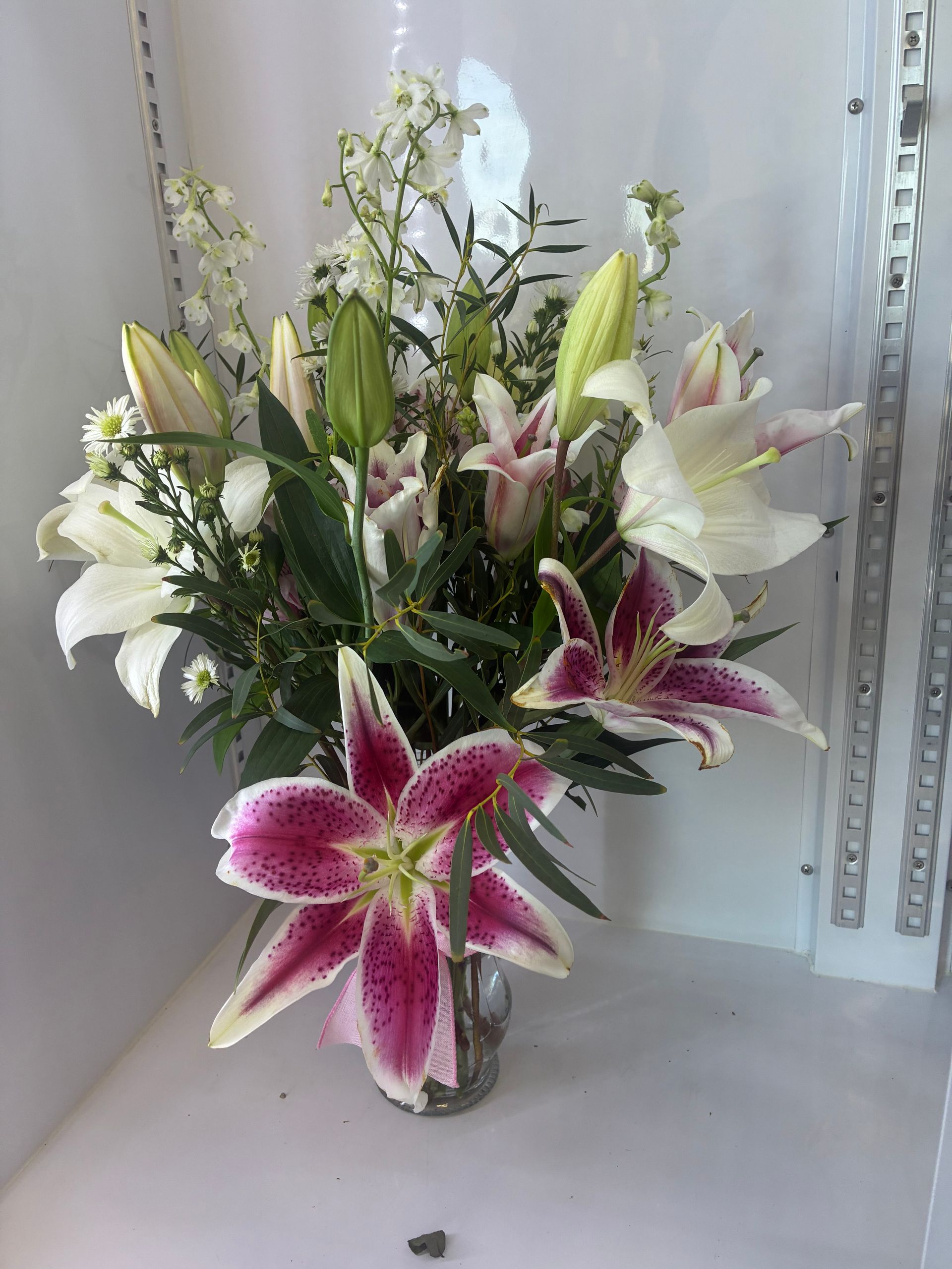 A bouquet of pink and white lilies with white filler flowers in a clear glass vase against a white wall.