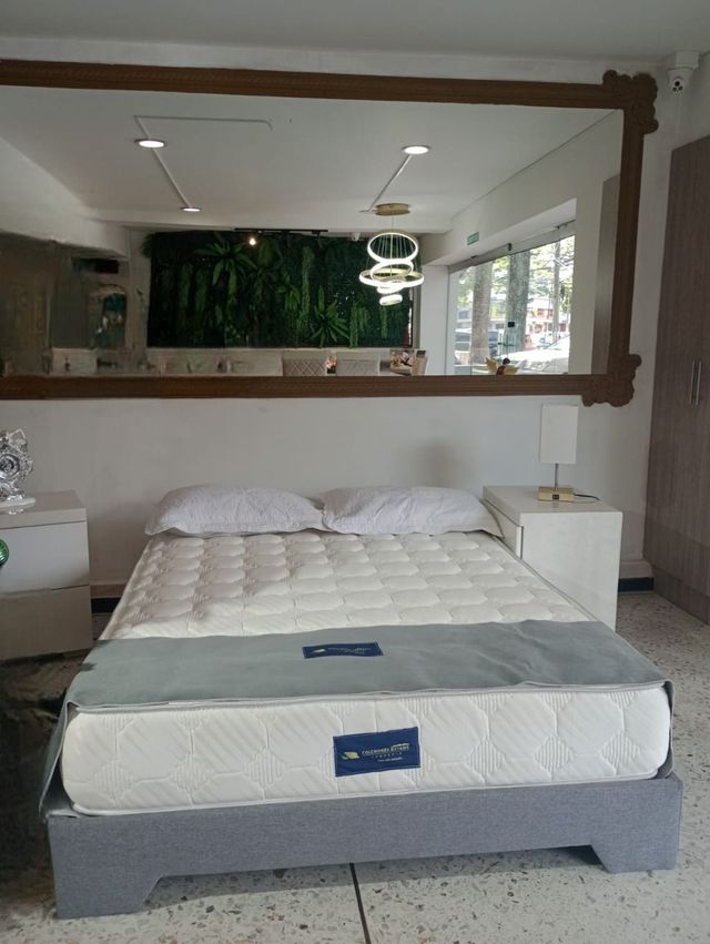 Dormitorio con la cama bien tendida frente a un gran espejo de pared y unas ventanas luminosas.