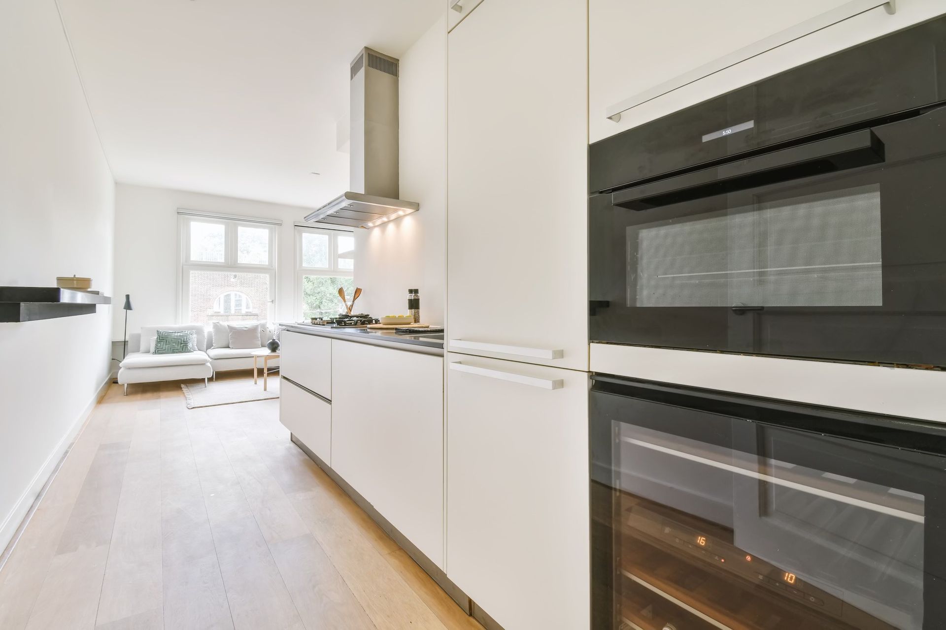Modern white kitchen with built-in oven and cabinets, leading to a living room with a window.