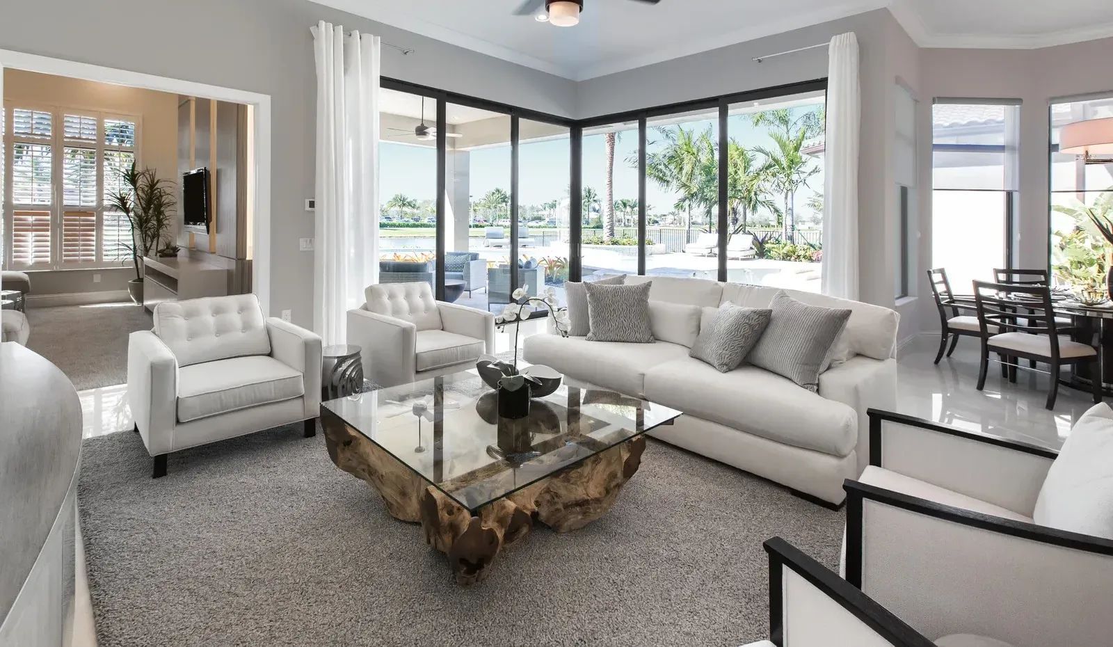 Bright modern living room with white sofas, glass coffee table, and large windows overlooking greenery