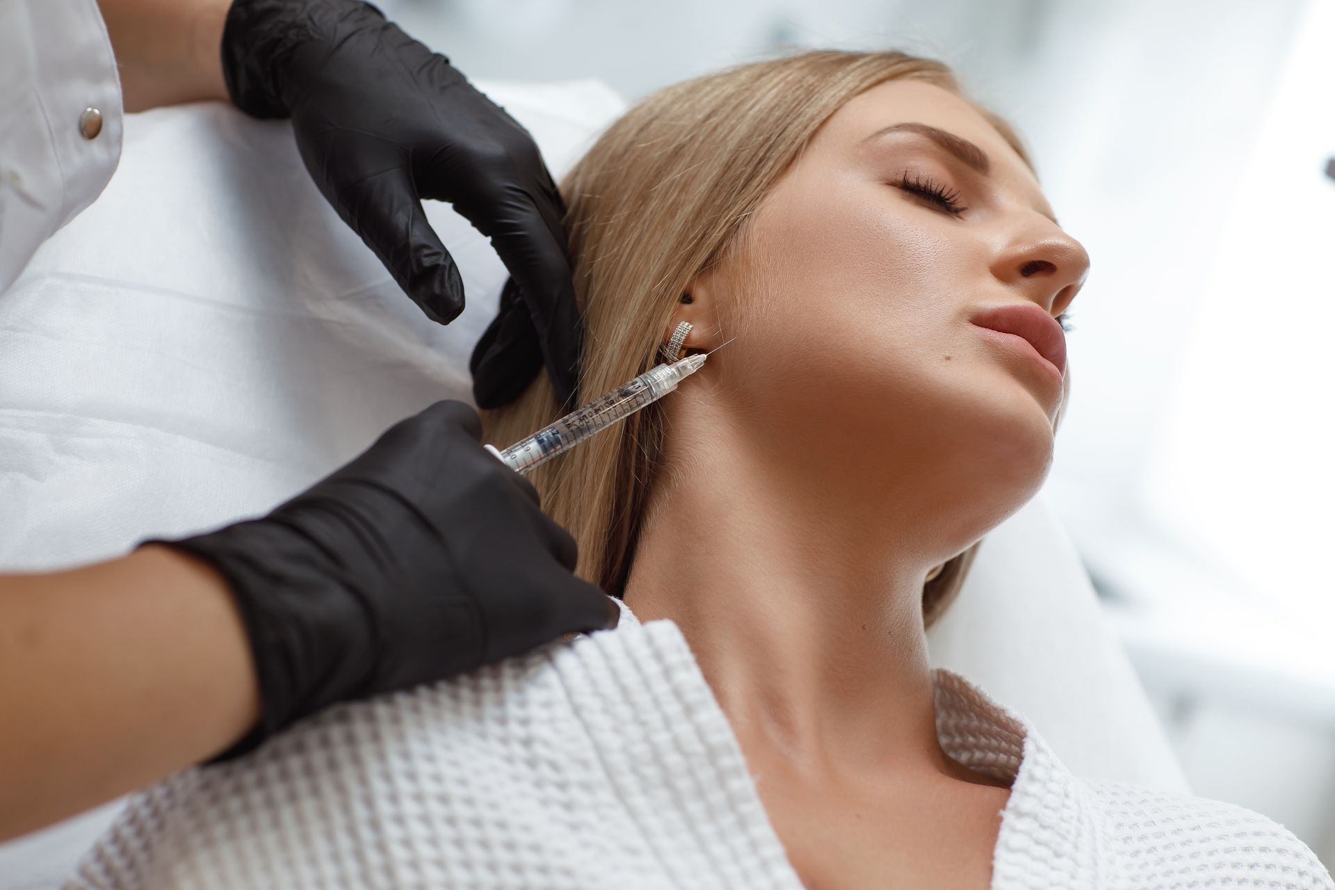 Gloved hands inject filler into a woman’s jawline in a clinical setting