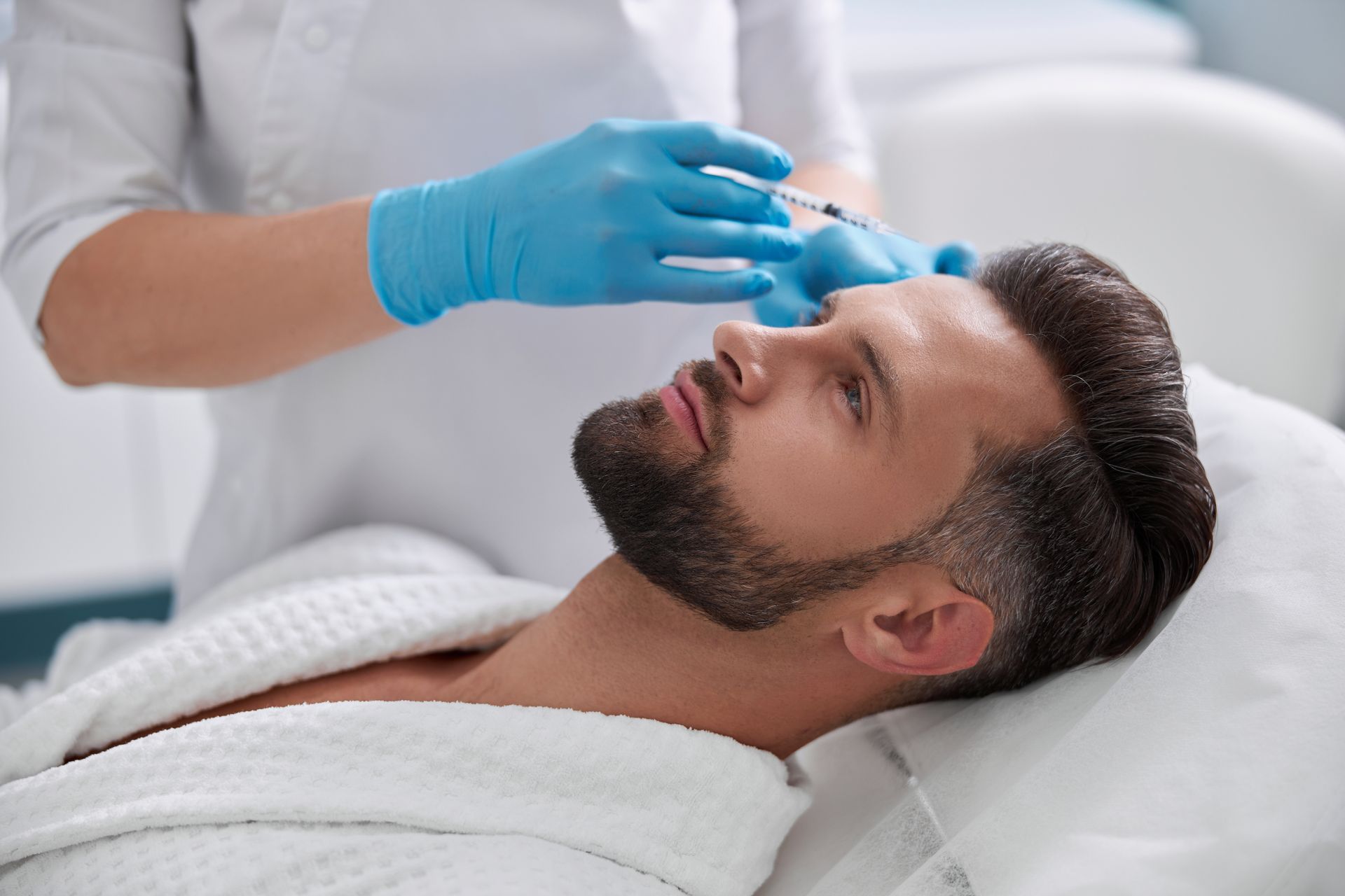 Gloved clinician applying a cosmetic injection to a man's forehead in a spa-like setting