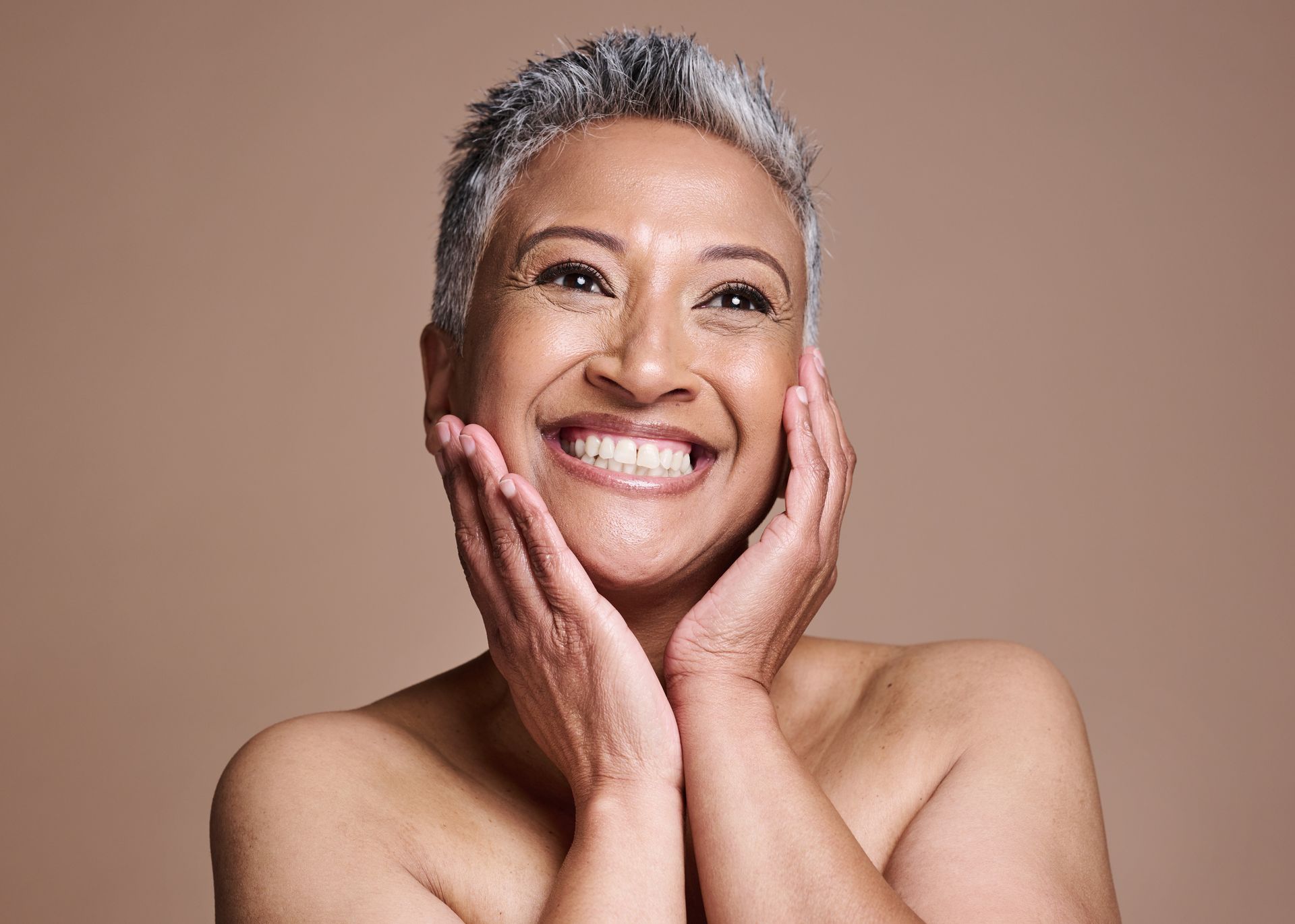 Woman with short gray hair smiles, touching her face, against a beige background.