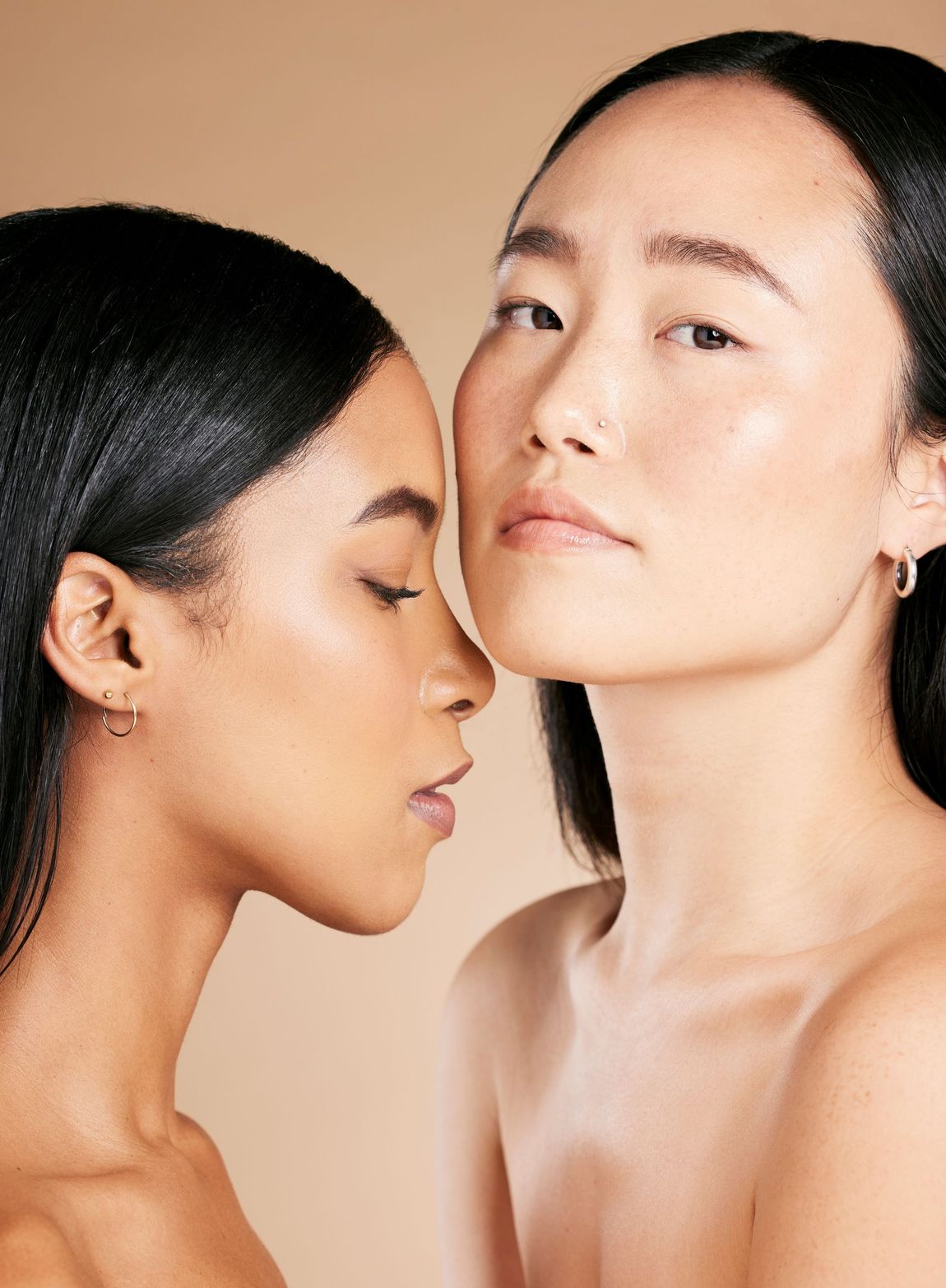Two women, with different skin tones, close together, looking towards camera and down with smooth skin, against neutral background.