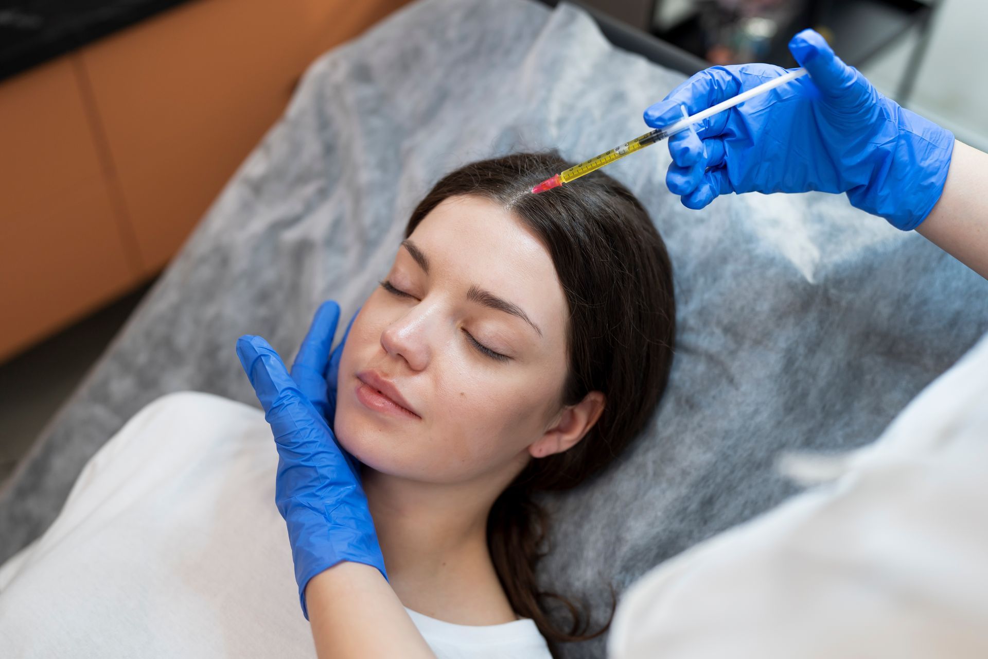 Aesthetic treatment being applied to a woman’s forehead with a syringe while she lies on a clinic bed.